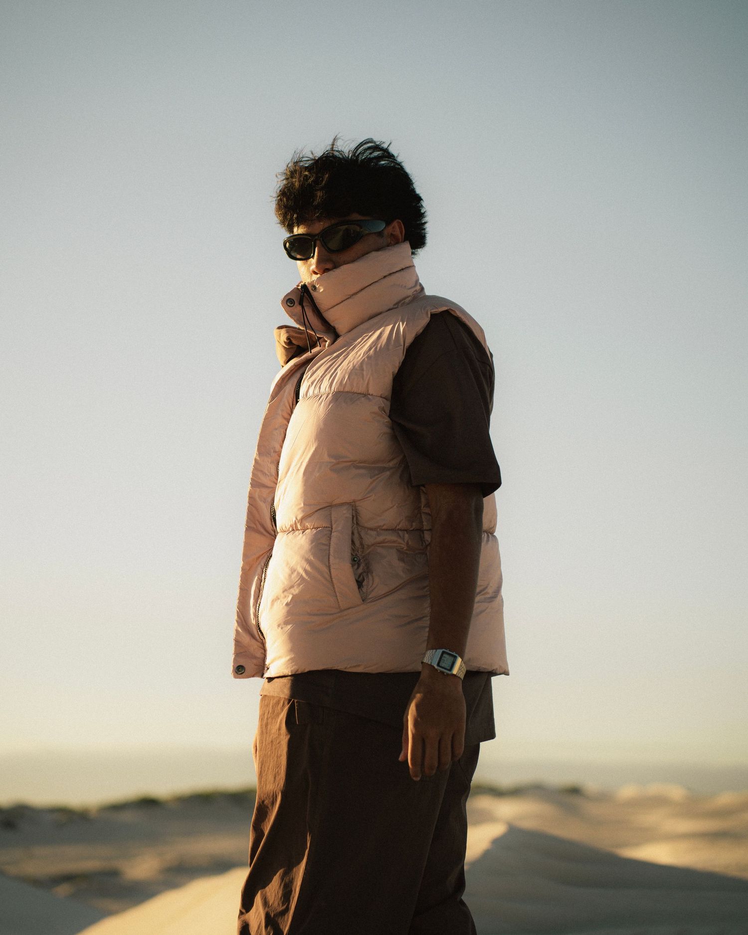 Person wearing a puffer vest and face covering standing in desert landscape at sunset.