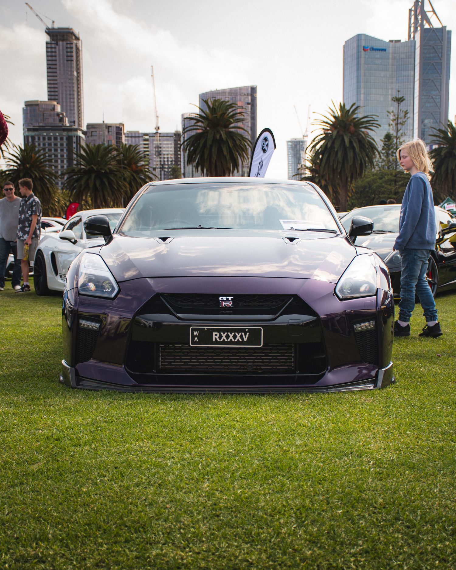 Purple Nissan GT-R displayed on grass at car show with city skyline in background.