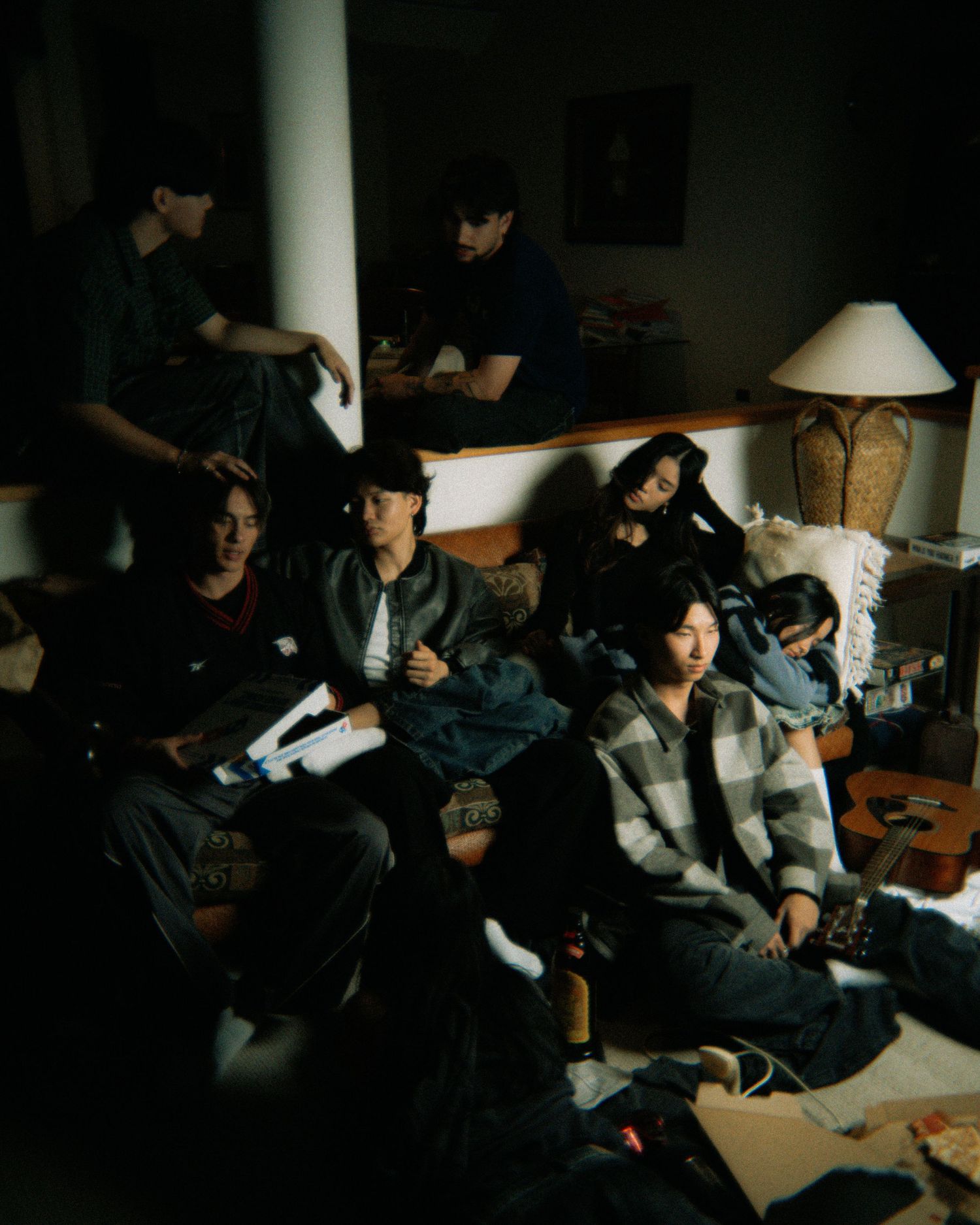 A dim-lit room with several people relaxing and hanging out around a table lamp on a casual evening.