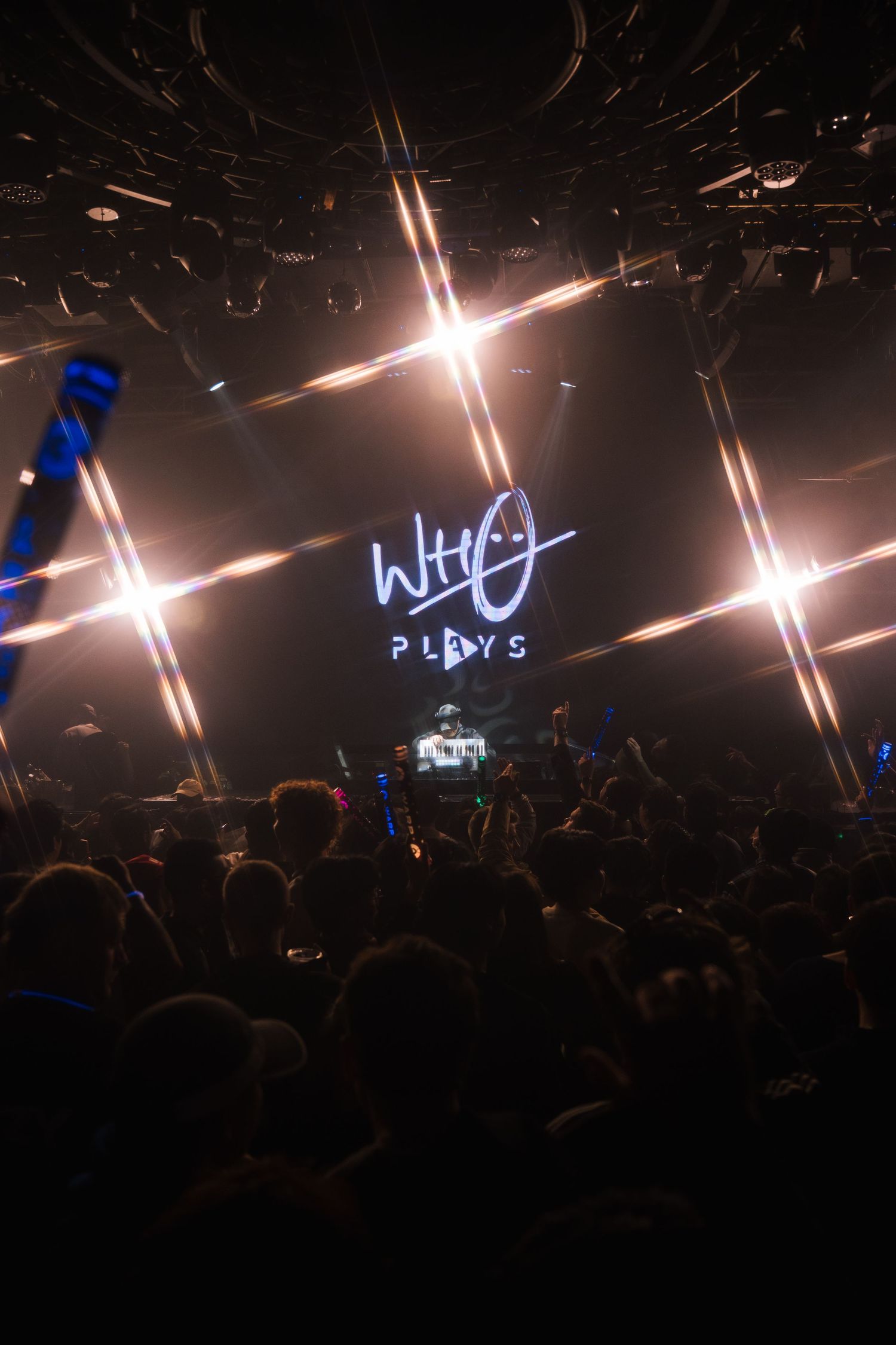 Concert venue with dramatic lighting beams cutting through dark atmosphere as crowd gathers for performance.
