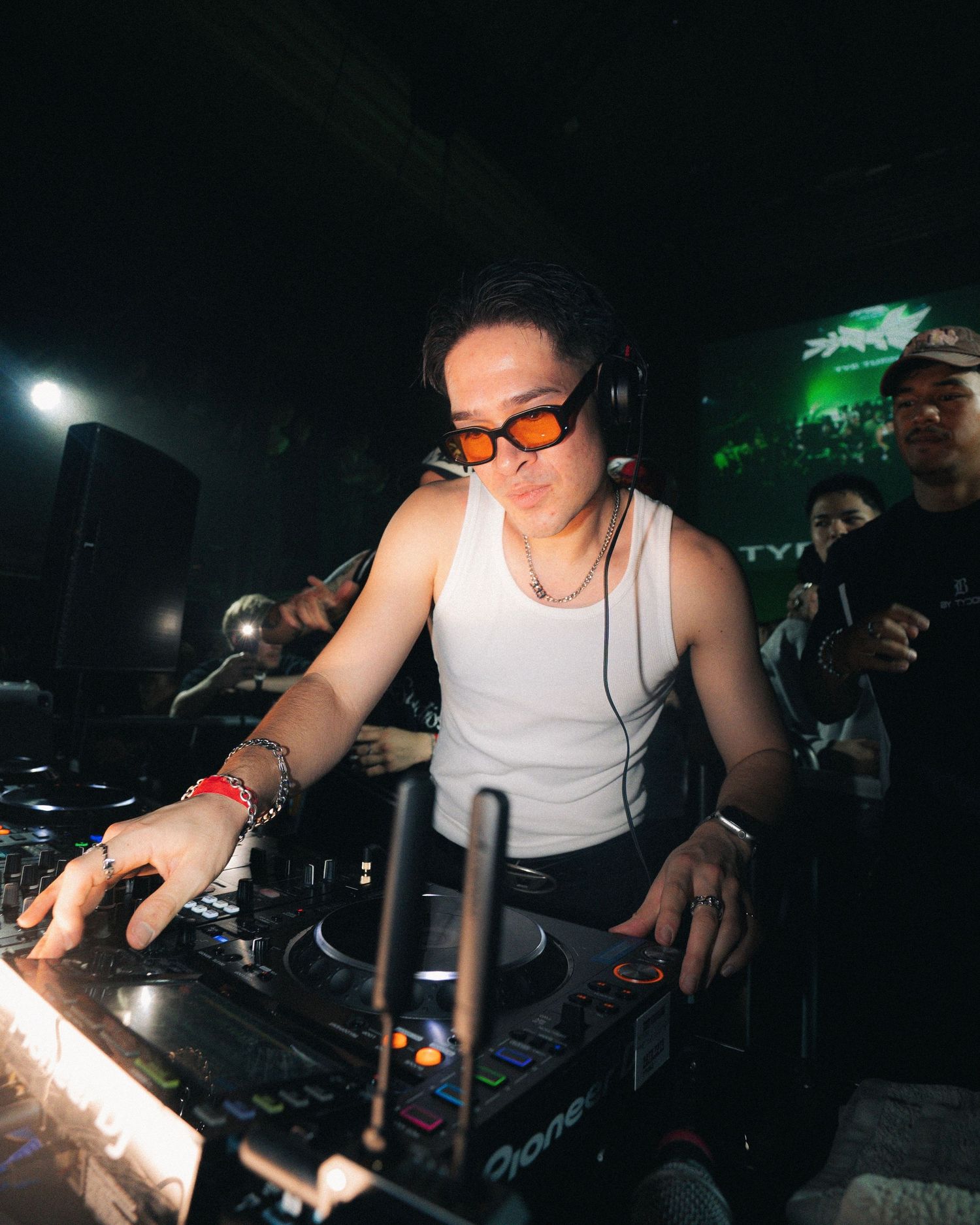 DJ in white tank top performs on Pioneer equipment in dark venue.
