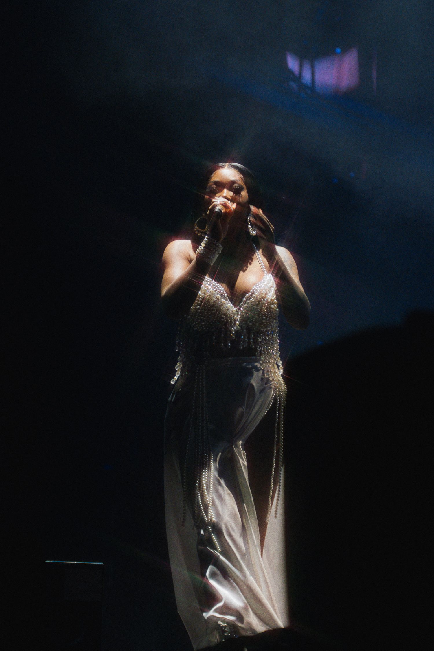 A singer in a sparkling white gown performs on a darkened stage with dramatic lighting.