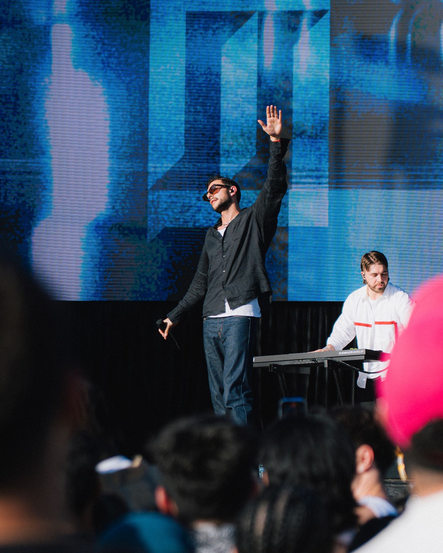 Performer stands with raised hand on stage during concert with blue lighting and keyboard player visible in background.