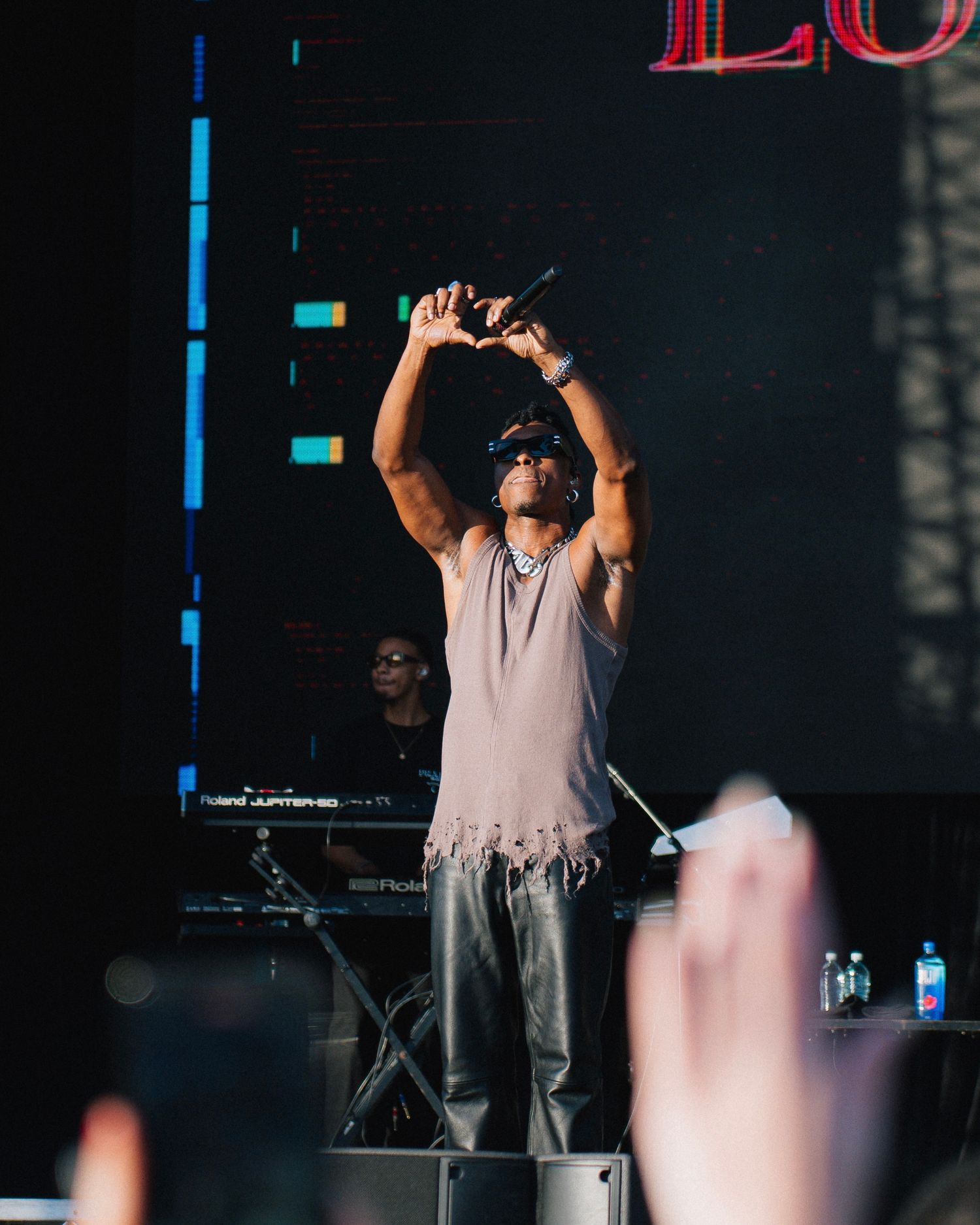 A performer in a sleeveless shirt raises their arms up while singing on stage during a concert.