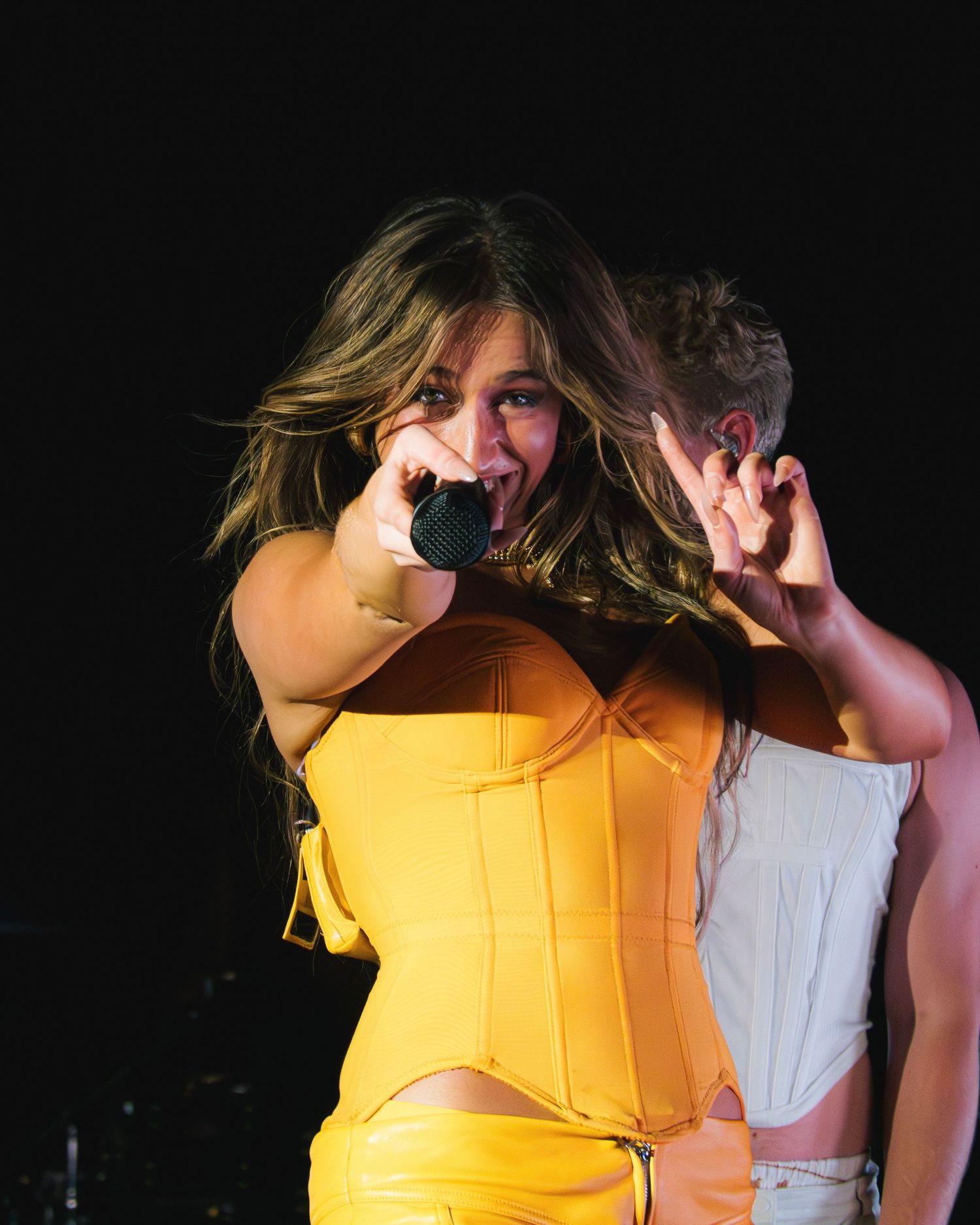 Energetic performer in yellow corset dress sings passionately on stage during dramatic concert performance.