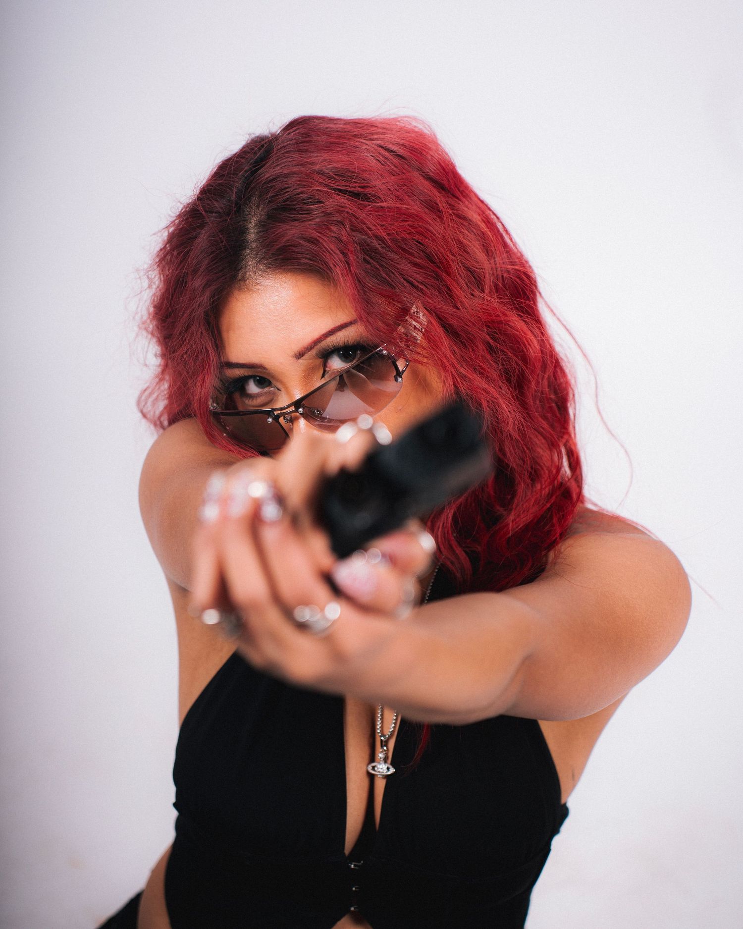 Person with bright red hair wearing black top aims a firearm while posing against white background.