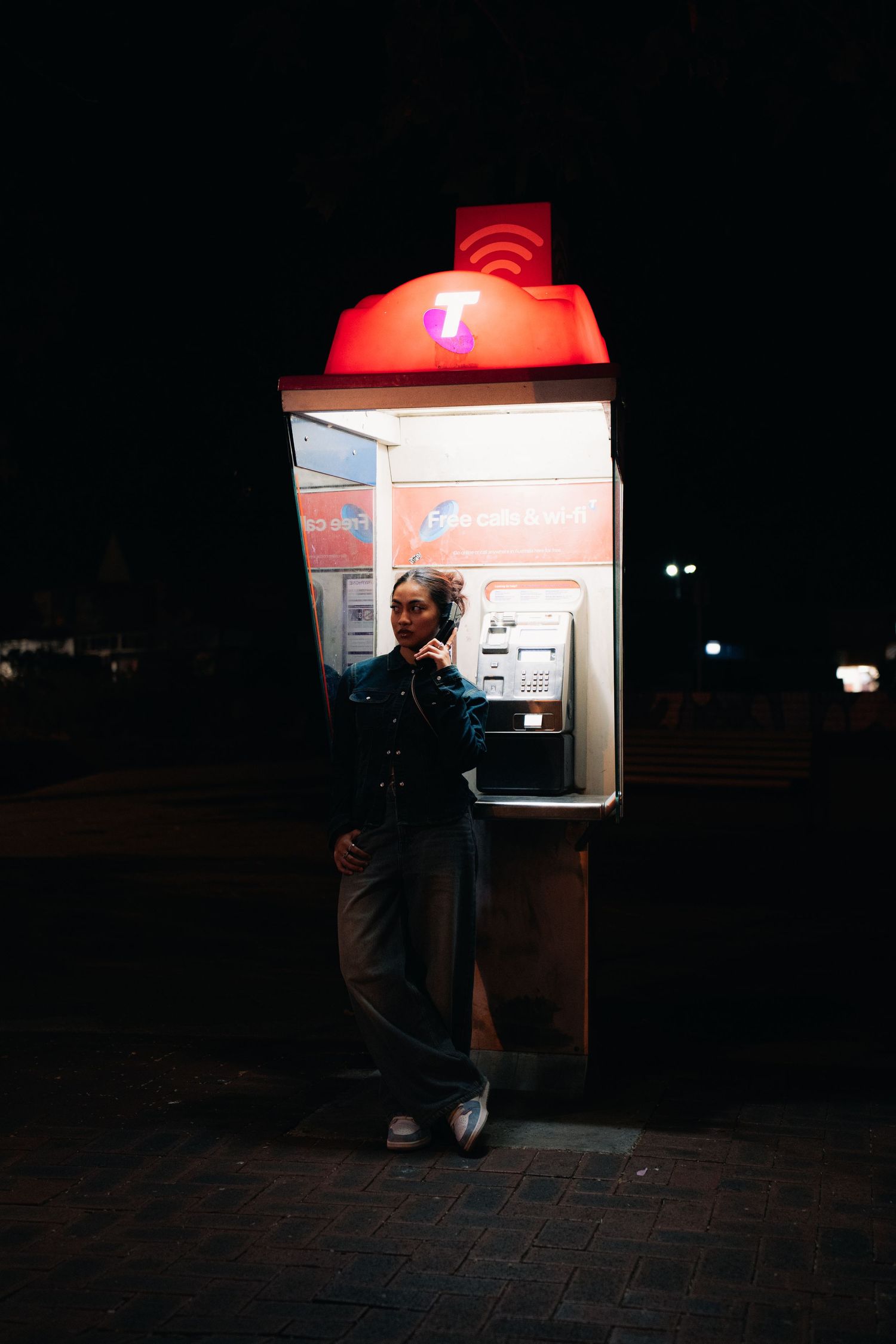 A silhouette stands at an illuminated phone booth with a red dome light at night on a brick sidewalk.
