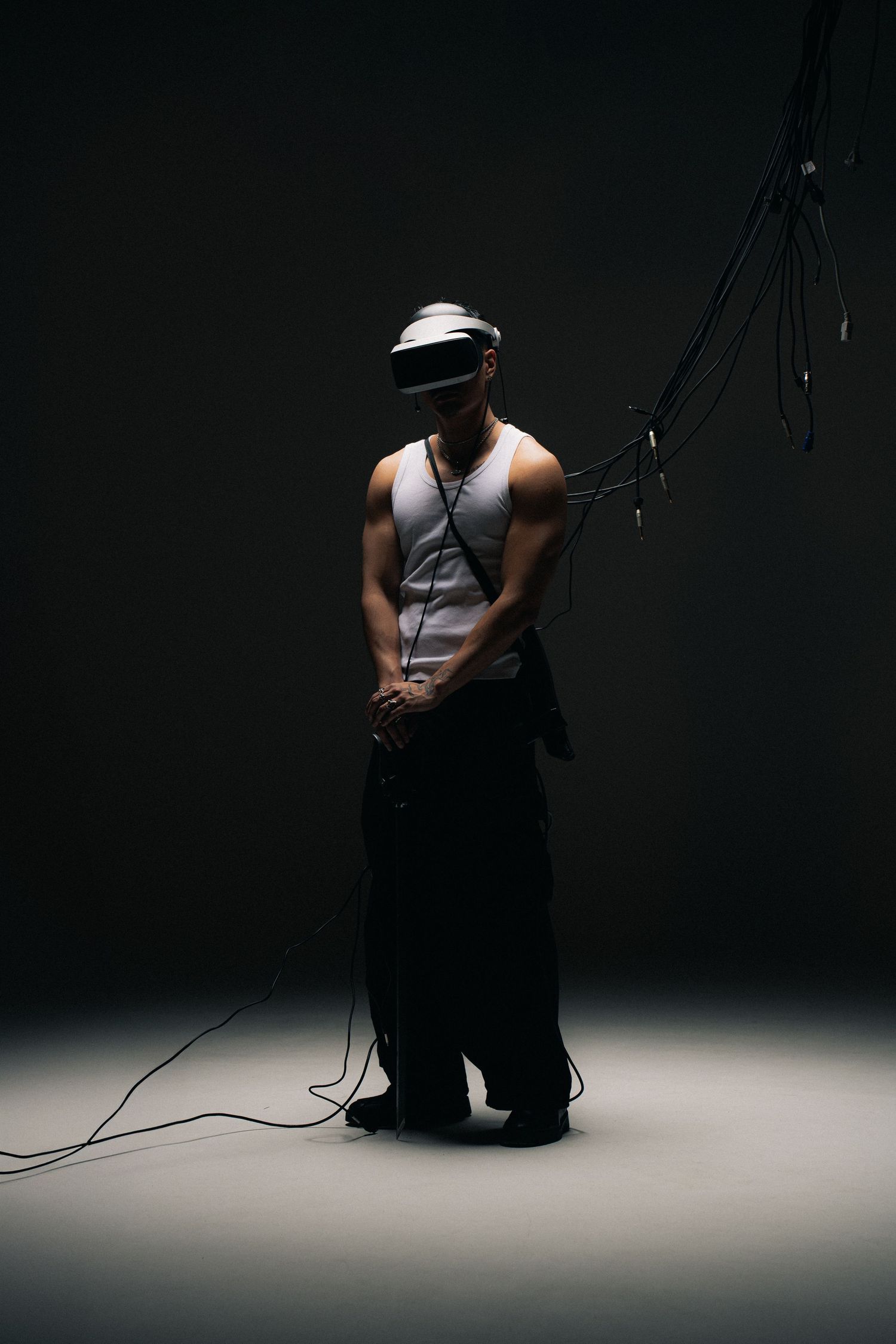 Dramatic low-key studio photo of person wearing VR headset and tank top standing in spotlight against dark background.