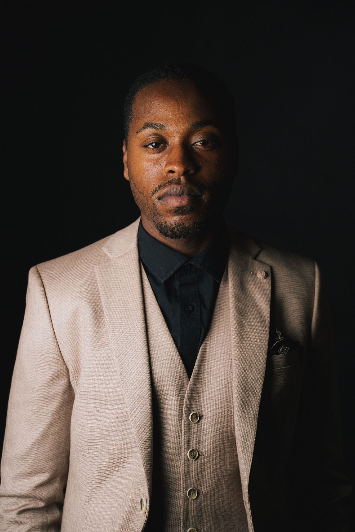 Professional studio portrait of someone wearing a beige three-piece suit with black shirt against dark background.