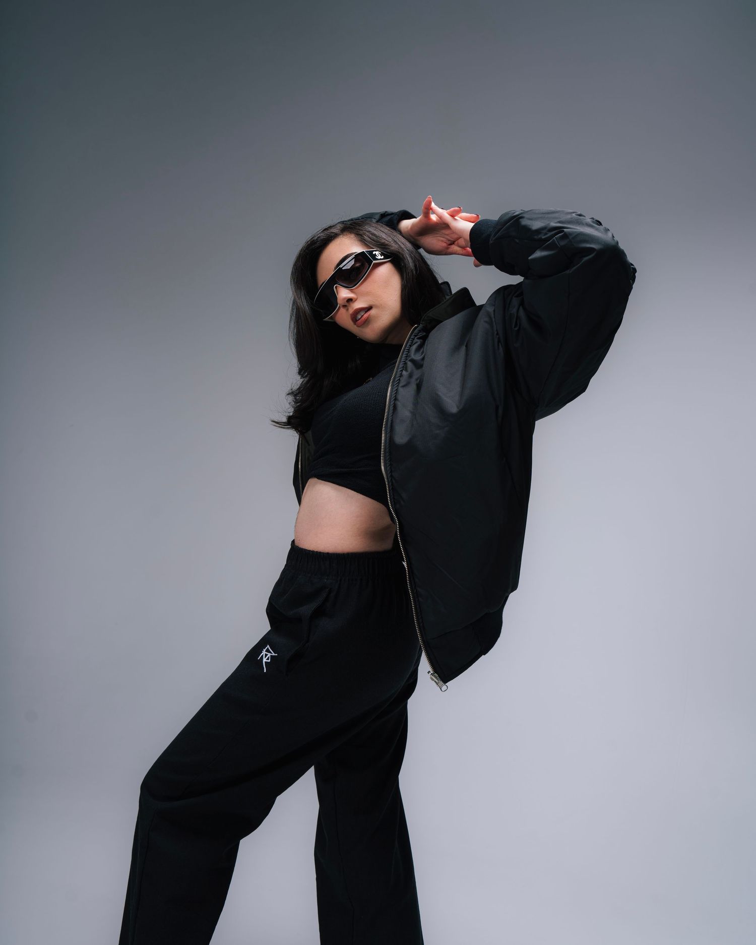 Person wearing sunglasses strikes dynamic pose in all-black streetwear outfit with bomber jacket and wide-leg pants.