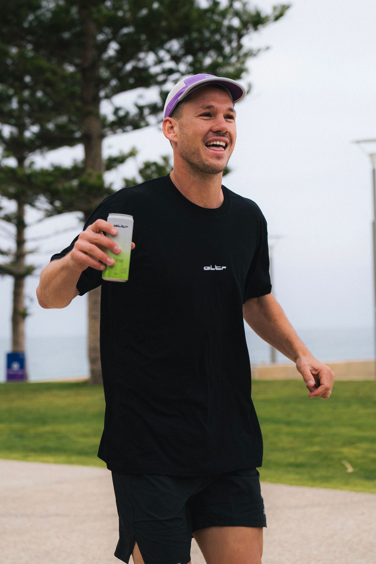 Someone wearing a black t-shirt and baseball cap enjoys a playful moment outdoors in a grassy coastal area.