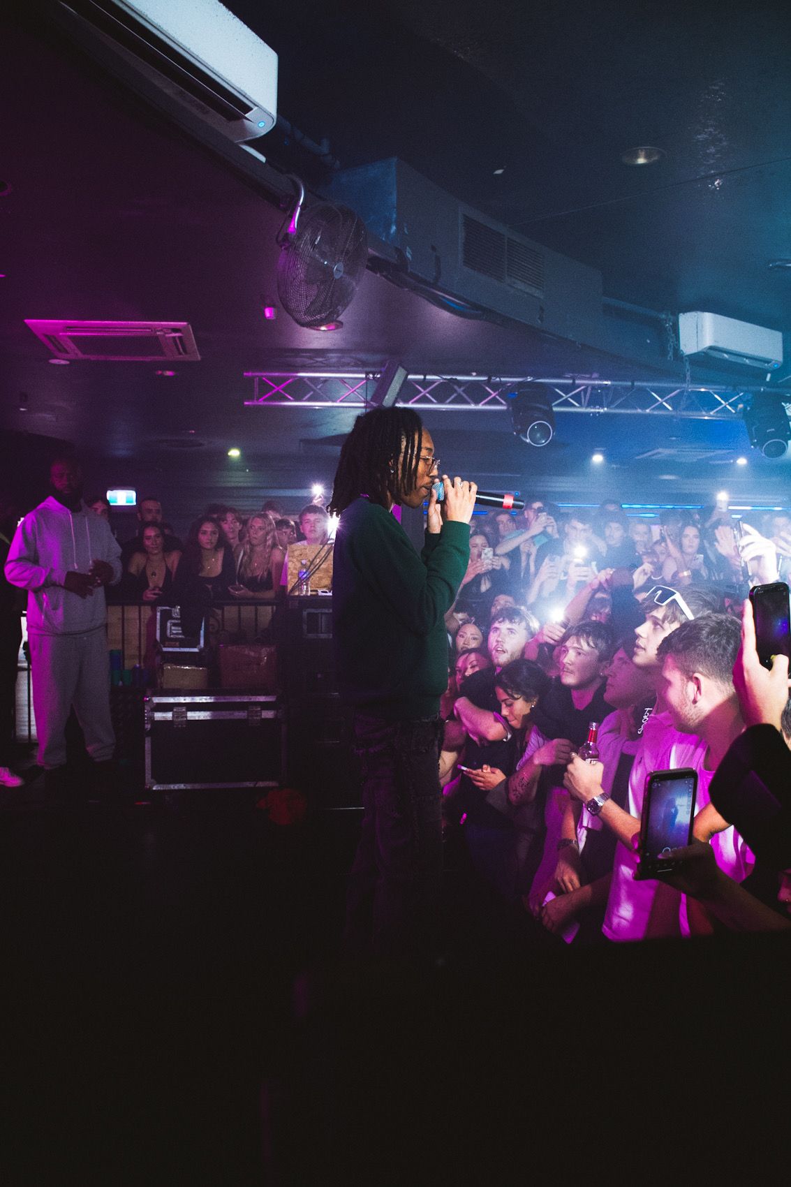 Concert crowd holding phones up as performer stands on stage with purple lighting effects.