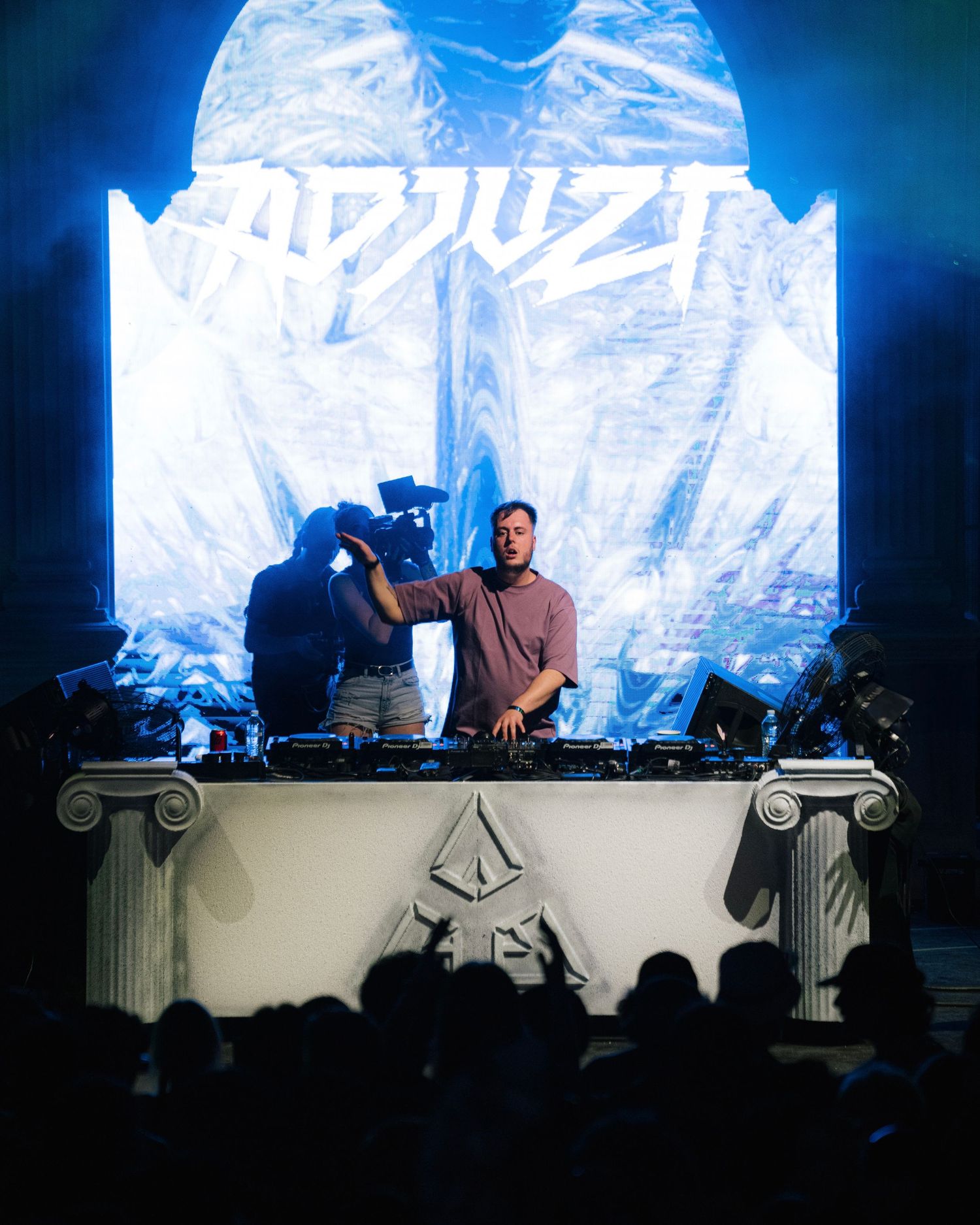 DJ setup with classical columns and glowing white backdrop at electronic music event.