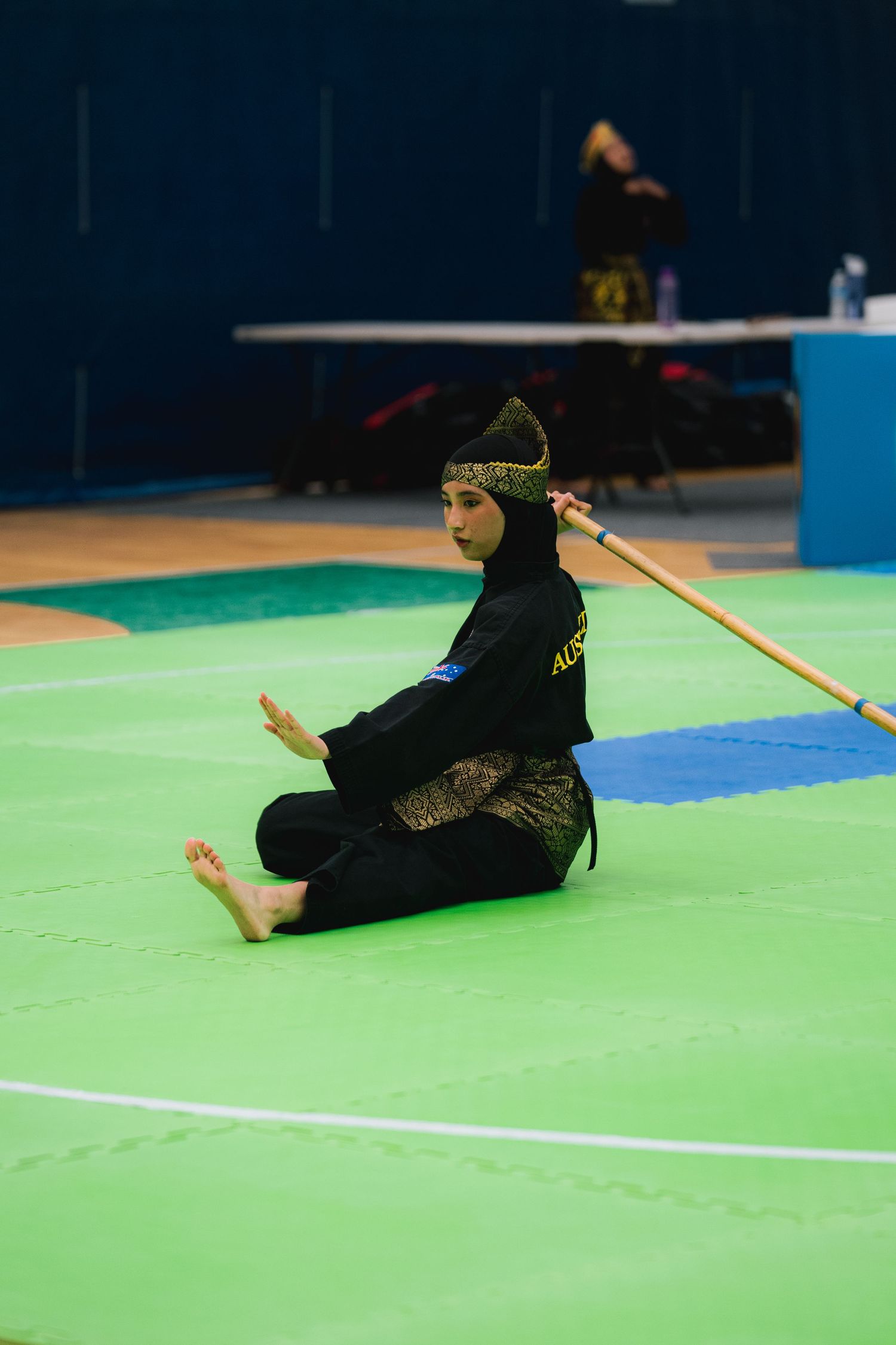 Martial artist in black uniform seated on green mat with staff weapon during competition.