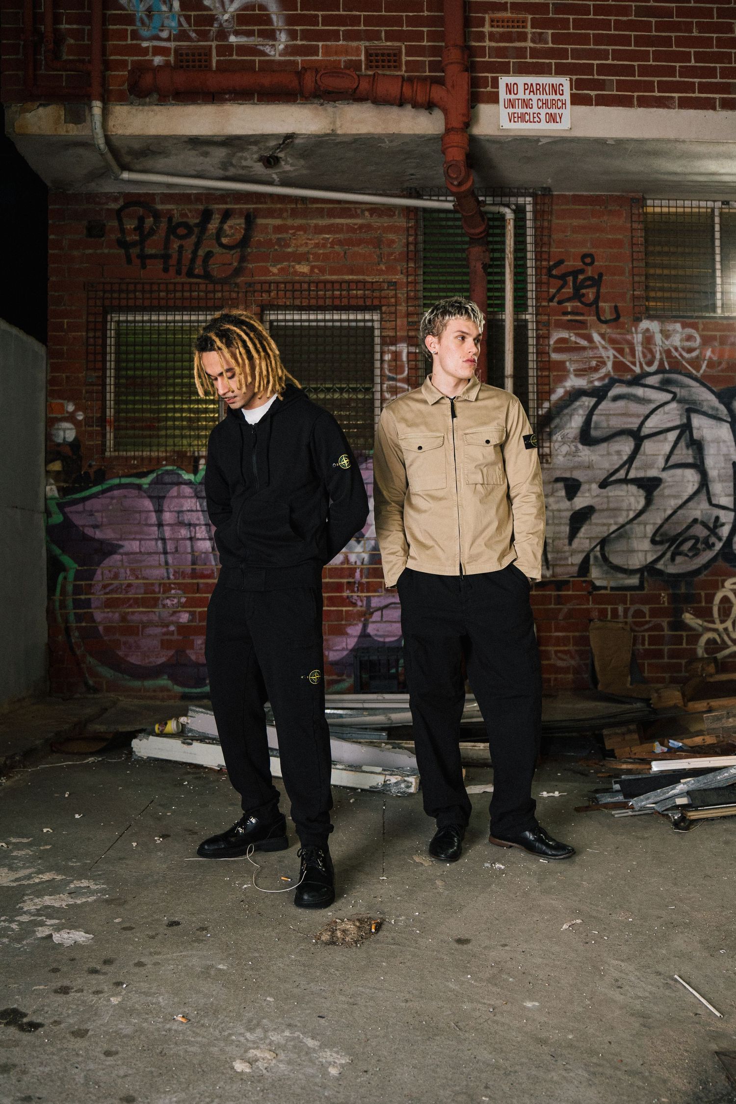 Two people pose against graffiti covered brick wall in urban setting.