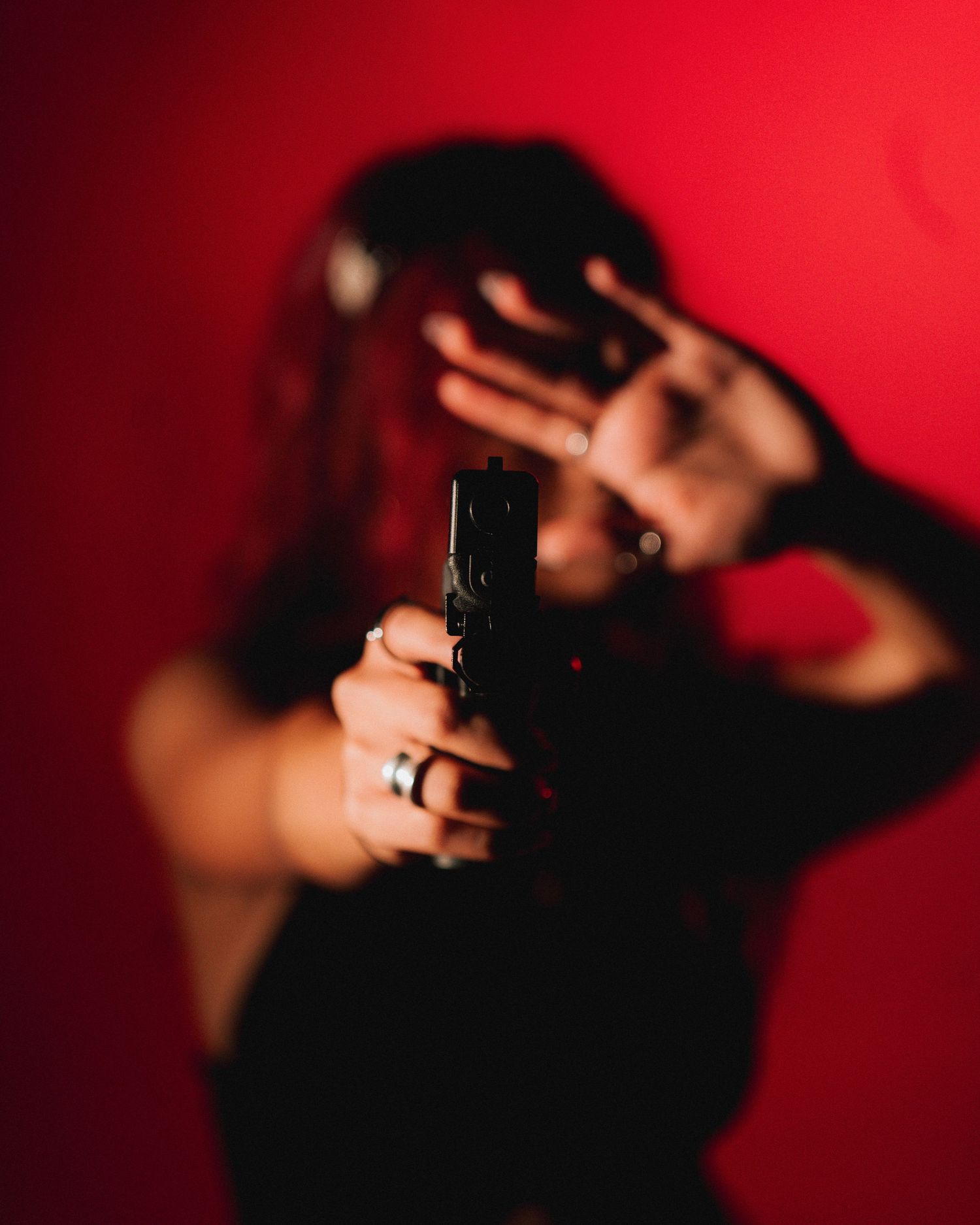 Person in black outfit holds a handgun while shielding face with hand against red background.