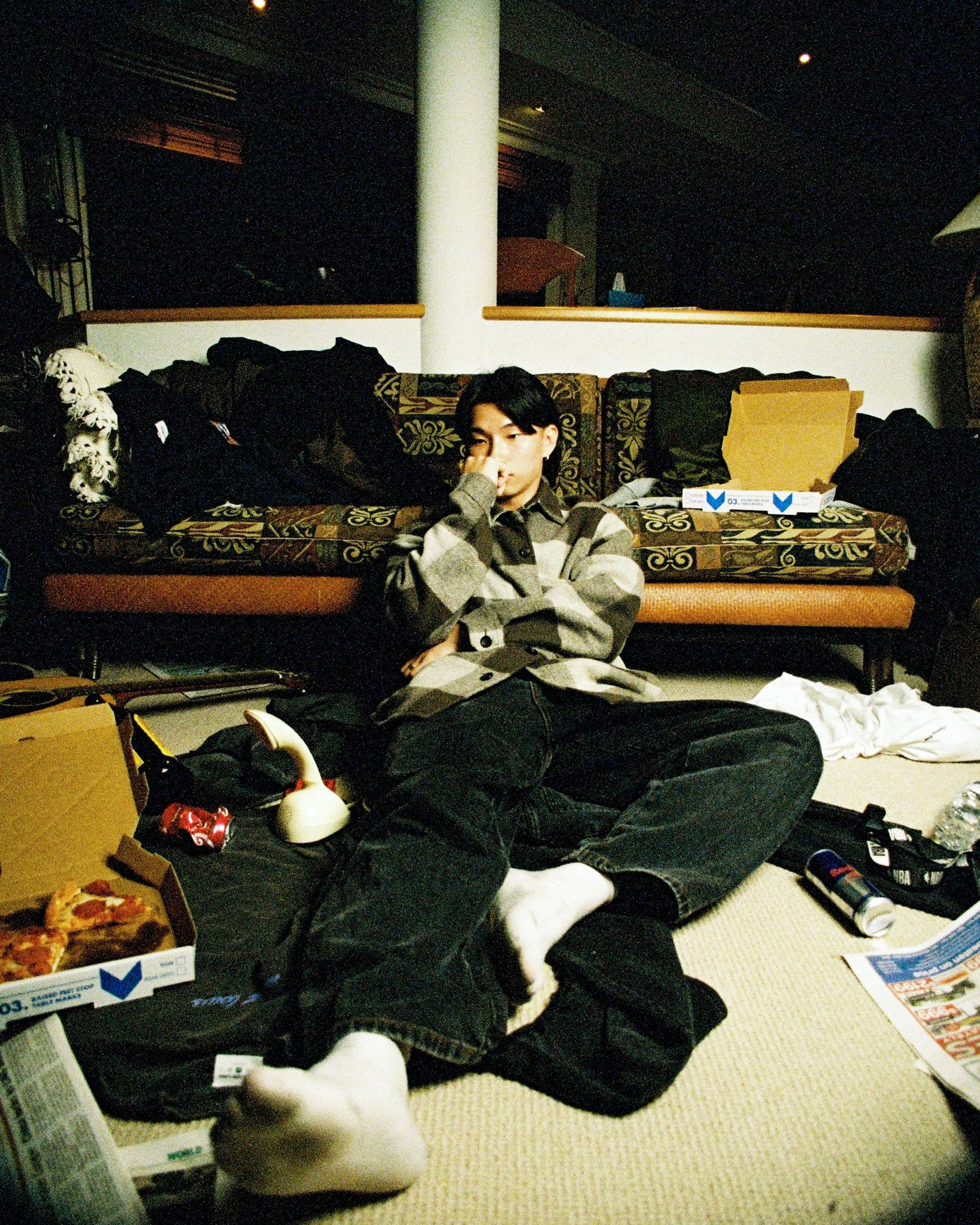 Black and white cats rest on messy couch with scattered clothes and pillows in dimly lit room.