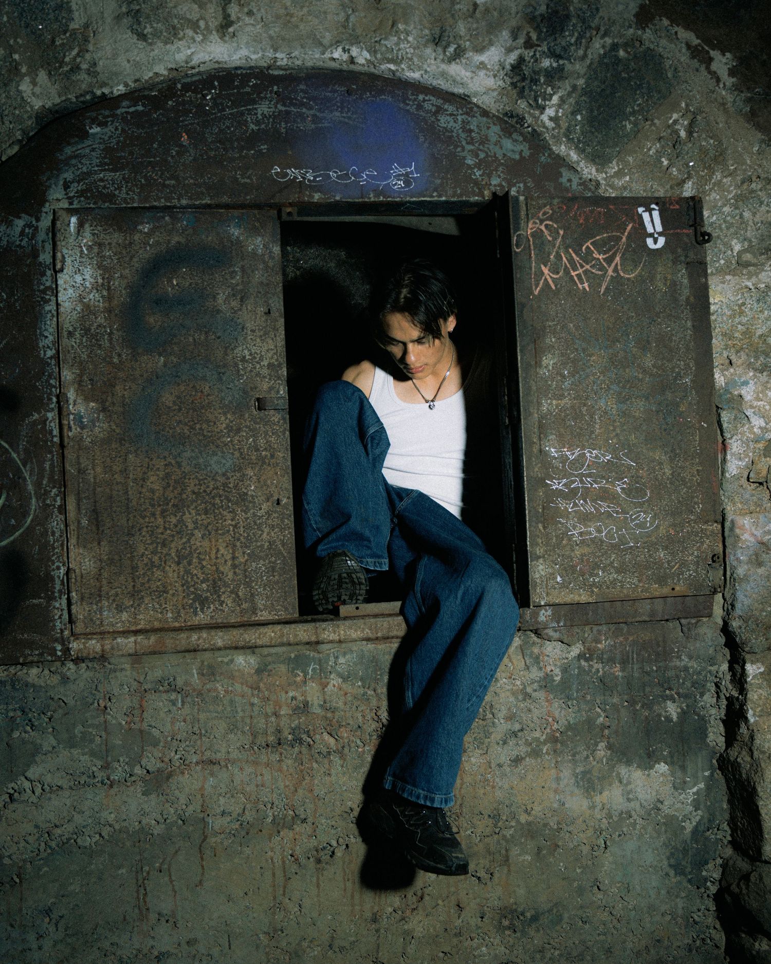 Figure in a white top and blue jeans perches in a dark stone archway with graffiti-marked walls.