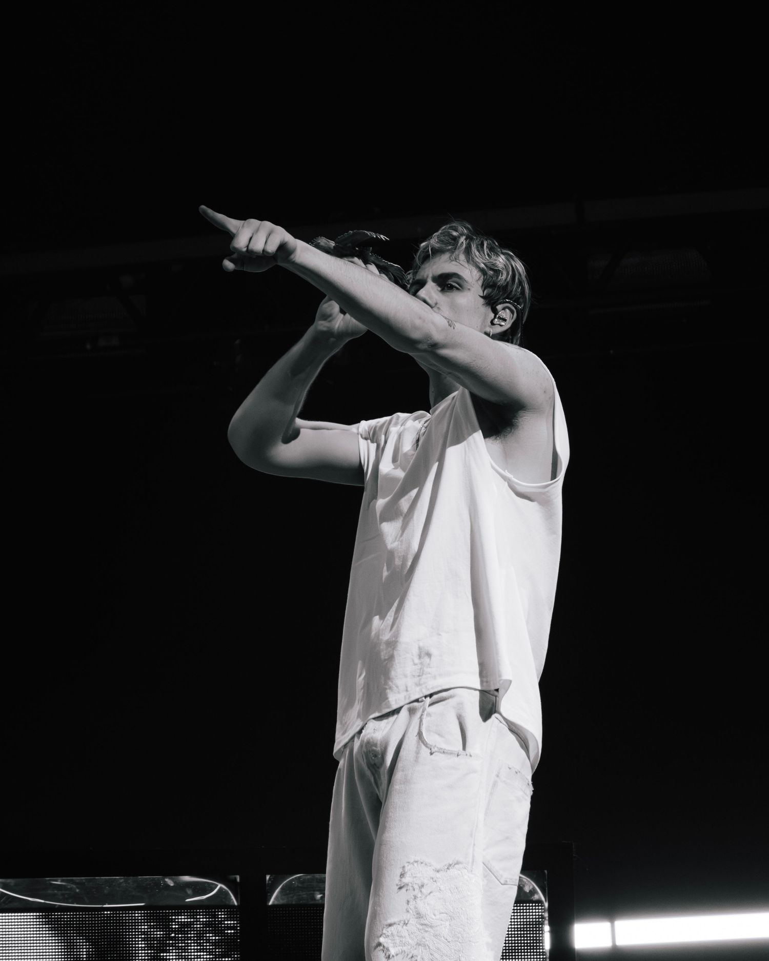 A performer in a white sleeveless shirt points upward while singing on stage in dramatic black and white lighting.