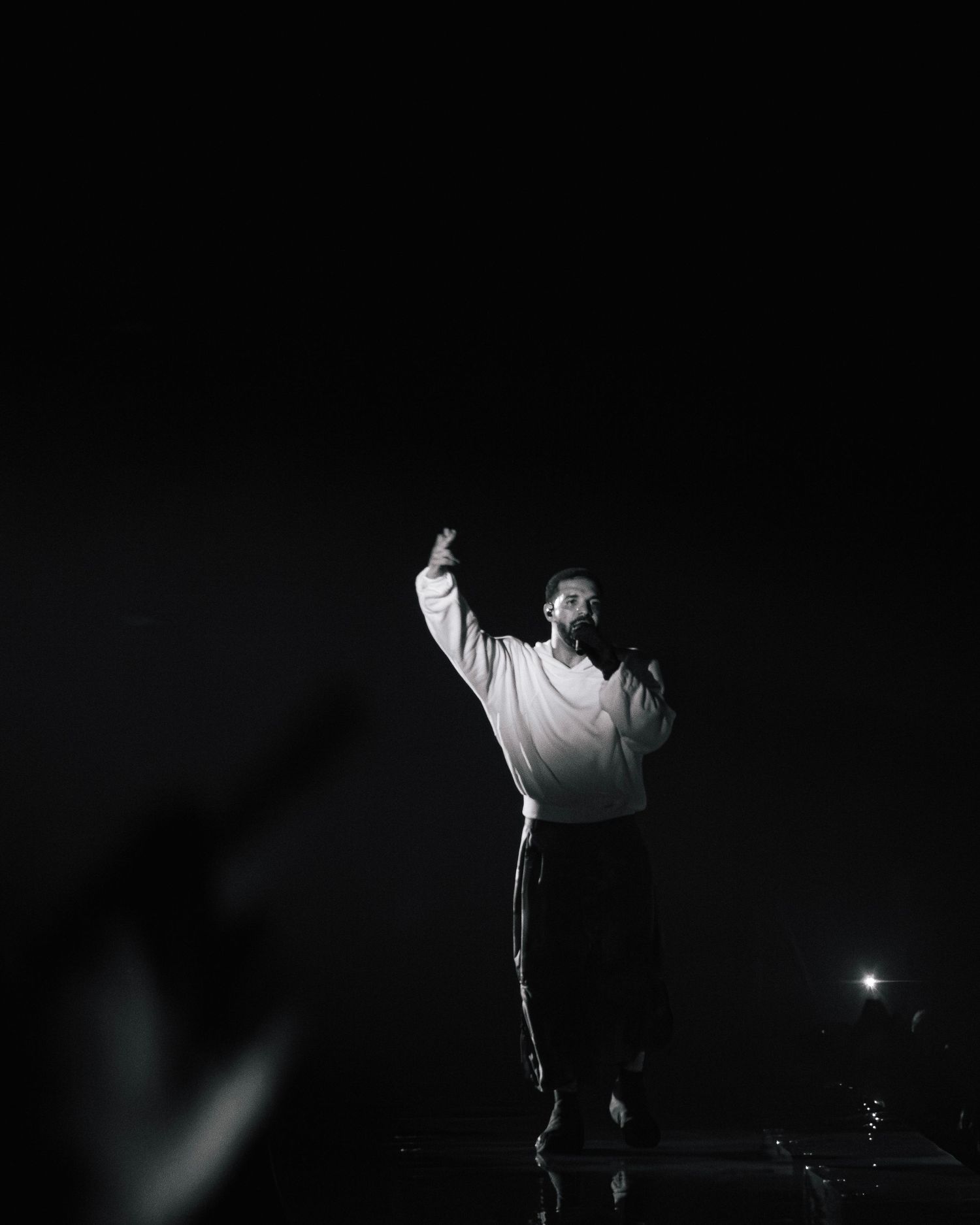 A performer in white shirt stands on stage in dramatic lighting, silhouetted against dark background with arm raised.