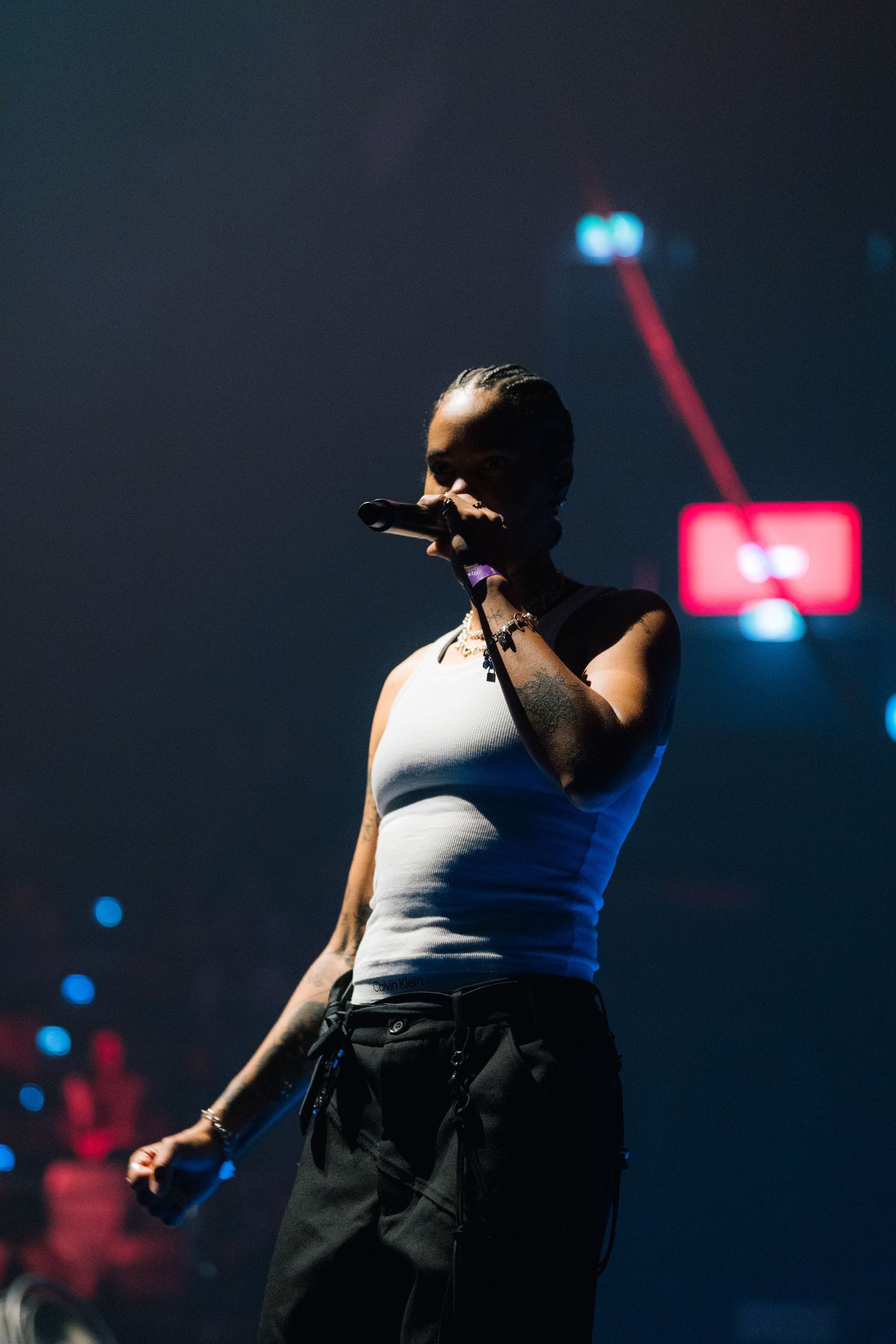 Performer in white tank top sings on dark stage with colorful lights and illuminated background.