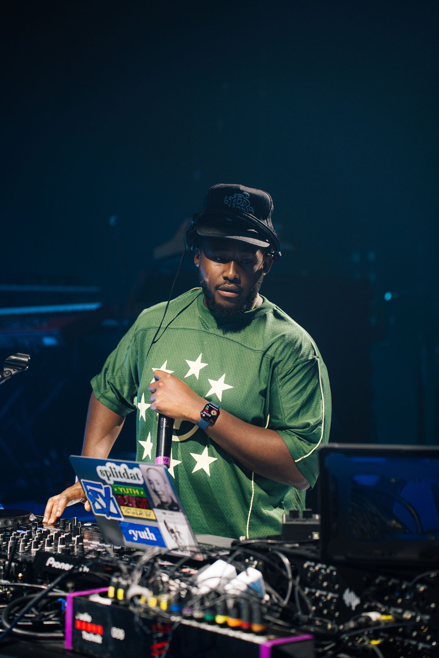 A DJ in a green star-patterned t-shirt and cap performs at a nightclub with professional mixing equipment.