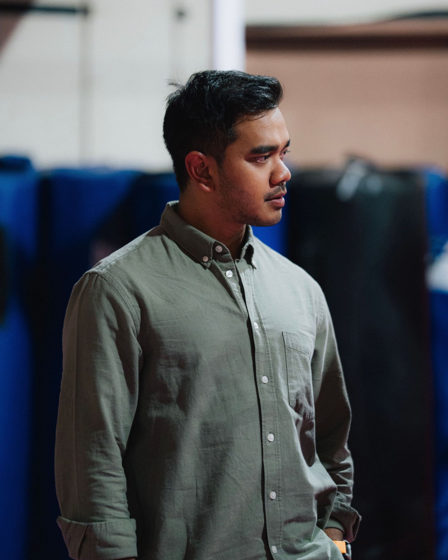 Portrait of person in olive button-down shirt against blue backdrop in indoor setting.