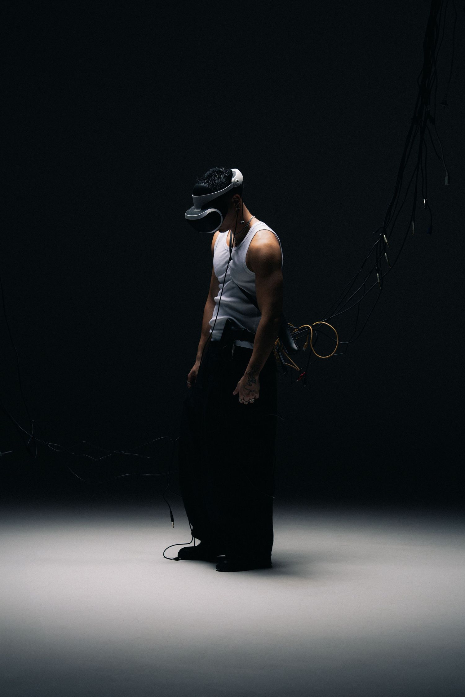 Person in VR headset and tank top standing in dramatic studio lighting with cables.
