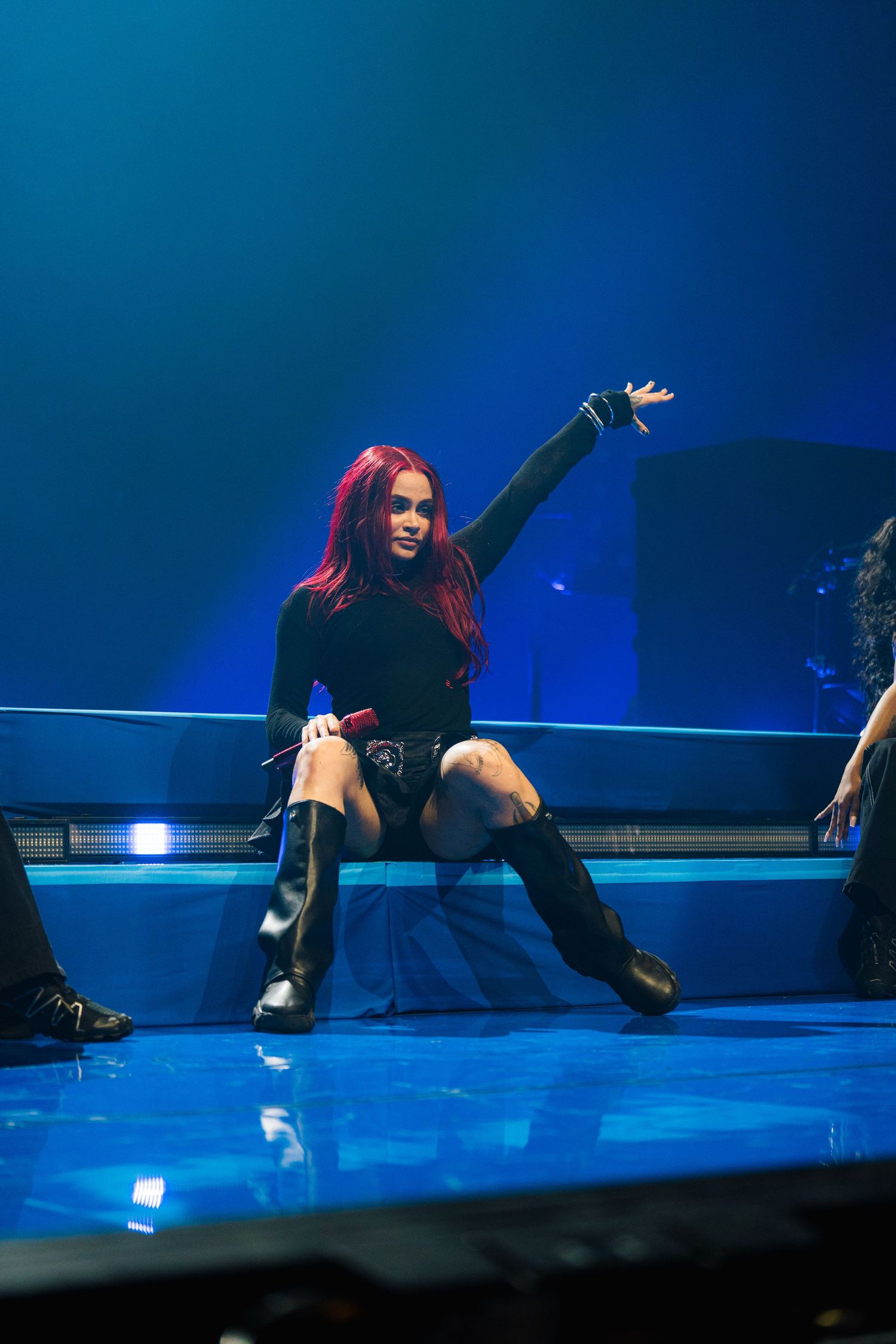 Performer with bright red hair in black outfit strikes dramatic pose on illuminated blue stage during live concert performance.