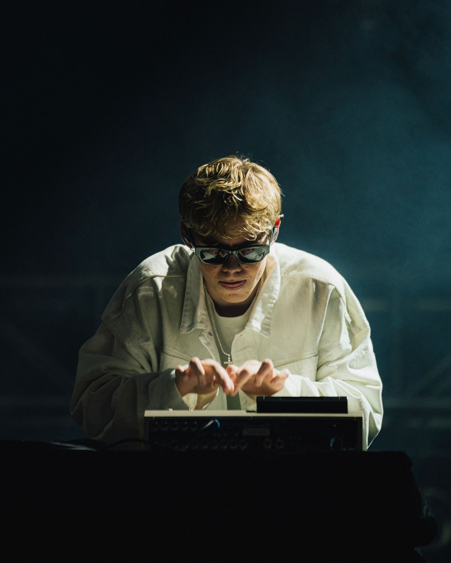 A person in a white outfit and eye mask types on a keyboard in a dark room with dramatic turquoise lighting.