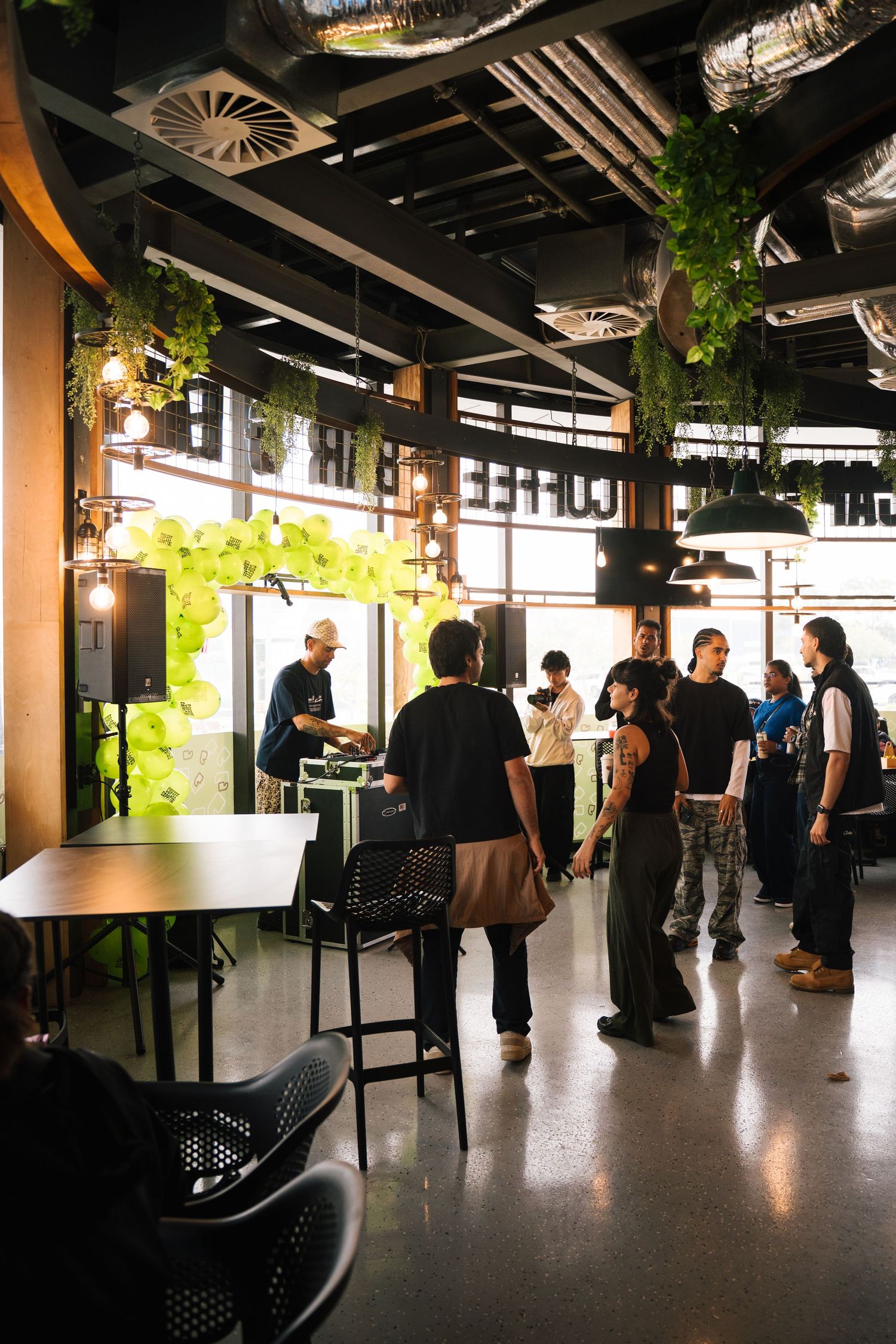 Modern restaurant interior with industrial ceiling, hanging plants and group of people gathered in casual dining space.