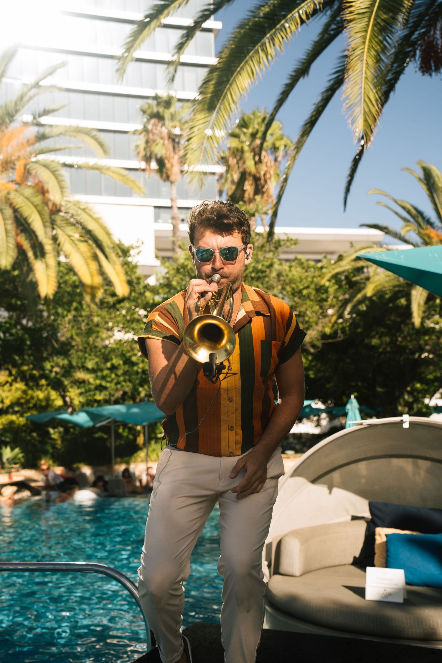 Person in brown jacket and gray pants lounging by luxurious resort pool with palm trees in tropical setting.