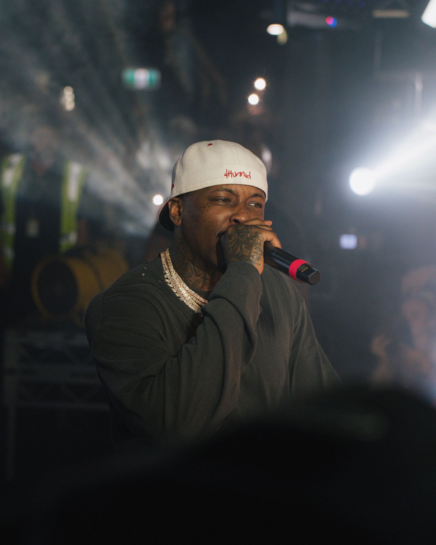 A performer in a white cap and black jacket holds a microphone while singing on stage with dramatic lighting in the background.