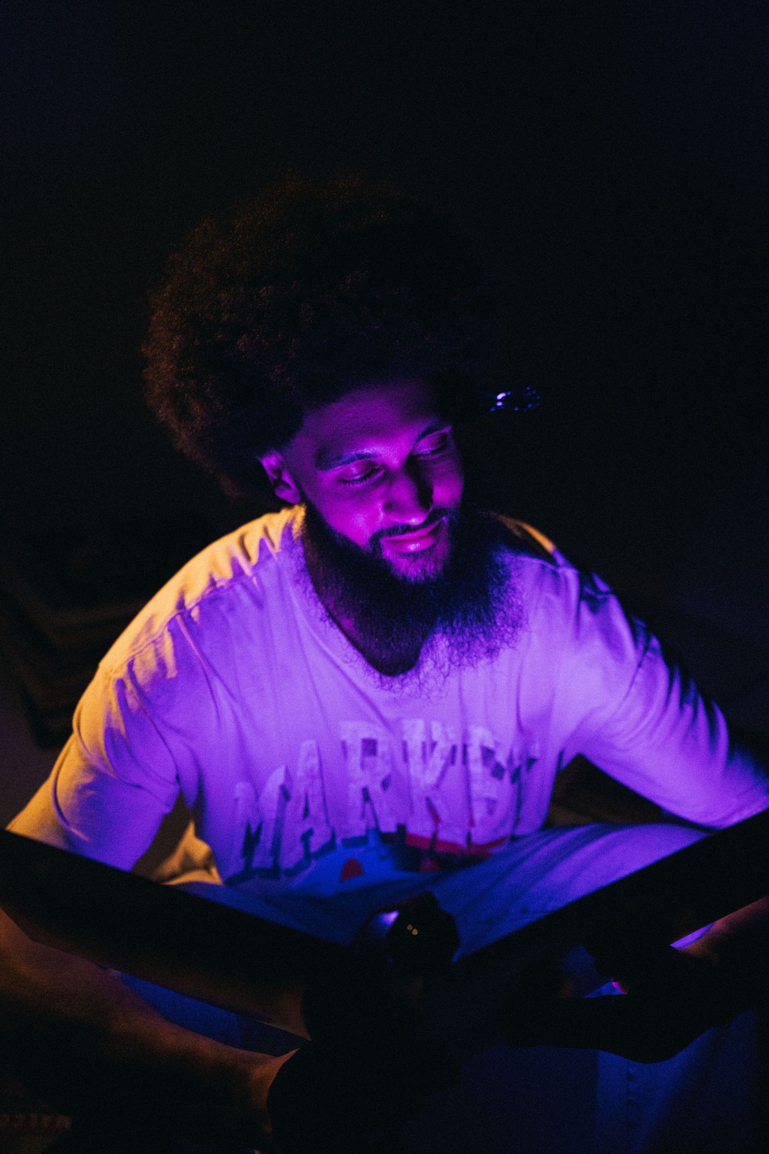 A person illuminated by purple lighting sits in a dark room with a warm yellow glow in the background.