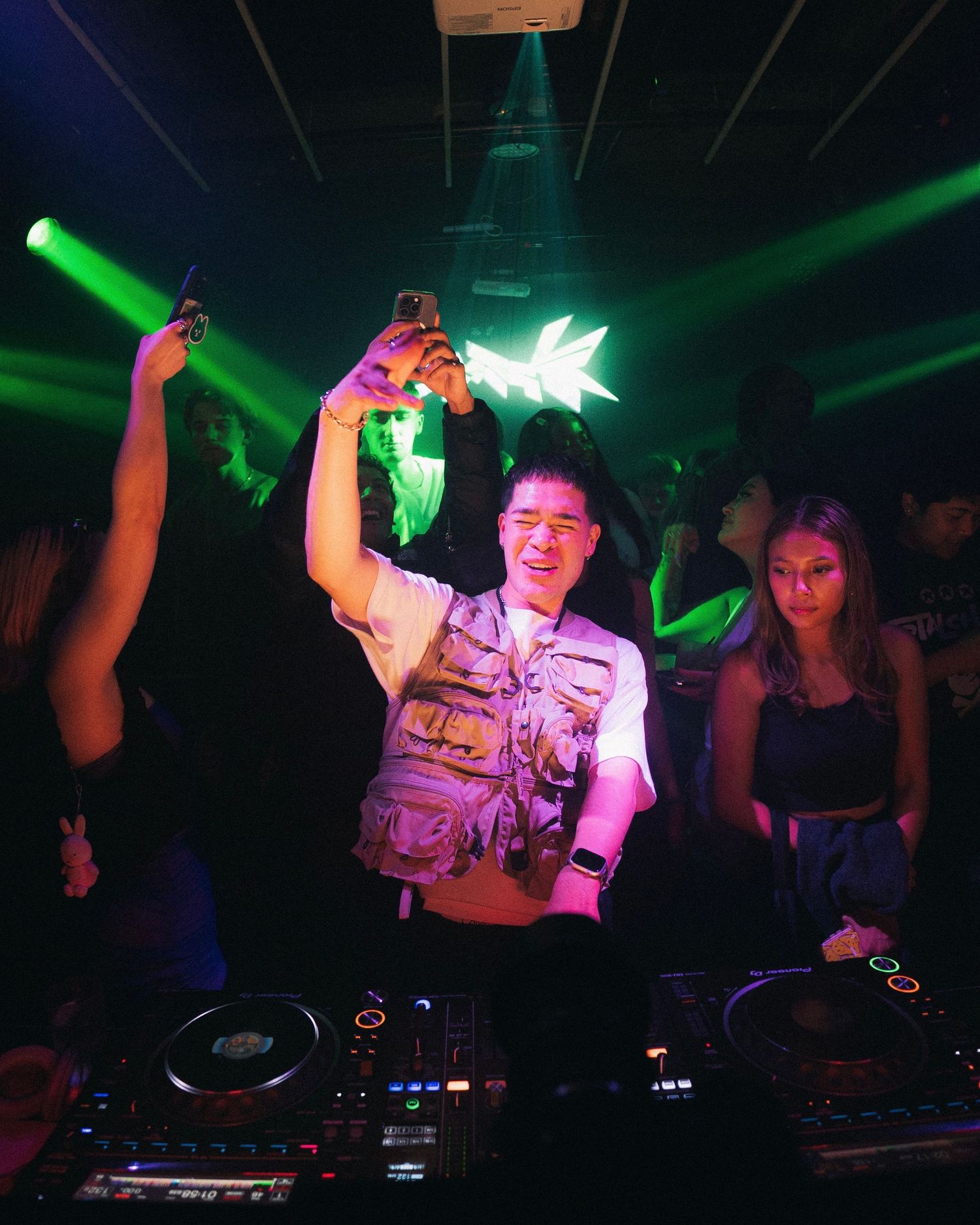 DJ performing at nightclub with green lighting effects and raised hands.
