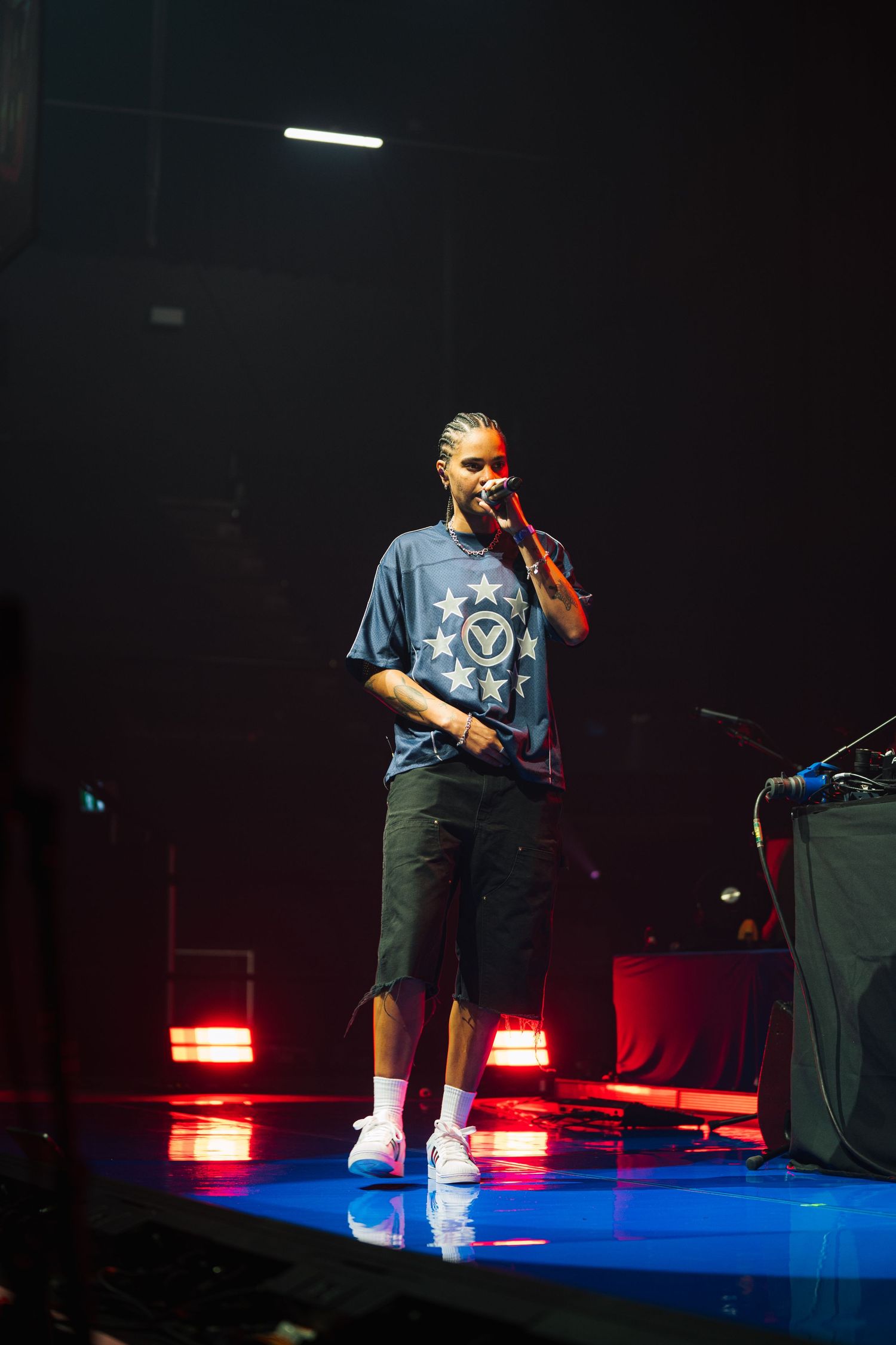 Performer in blue t-shirt and shorts stands on stage with dramatic lighting and red accent lights during concert performance.