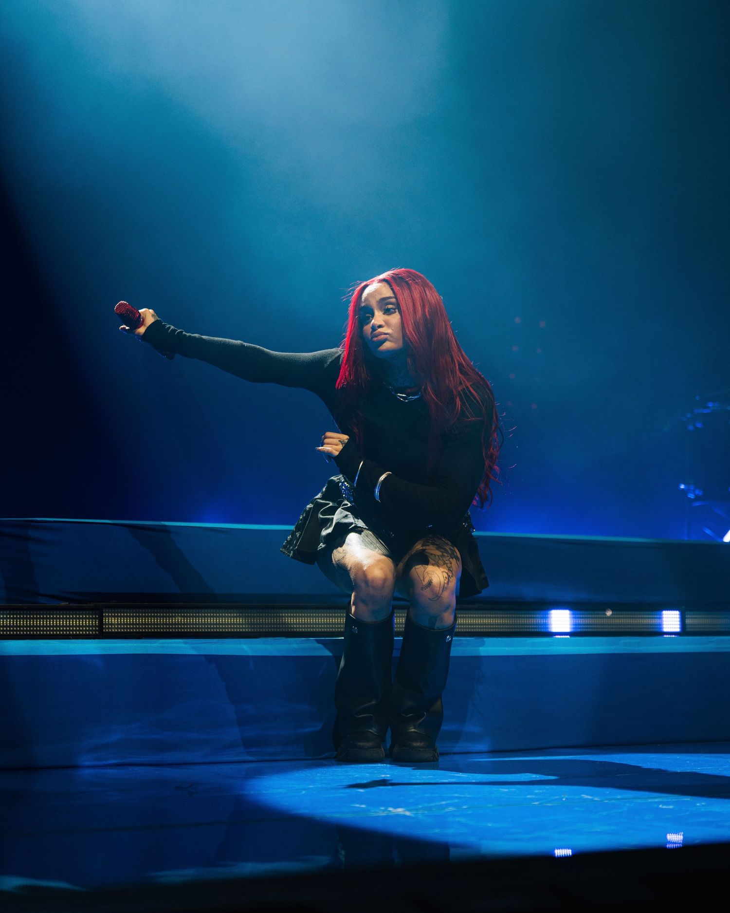 A performer with red hair in a black outfit strikes a dramatic pose on a dimly lit blue stage during a concert performance.