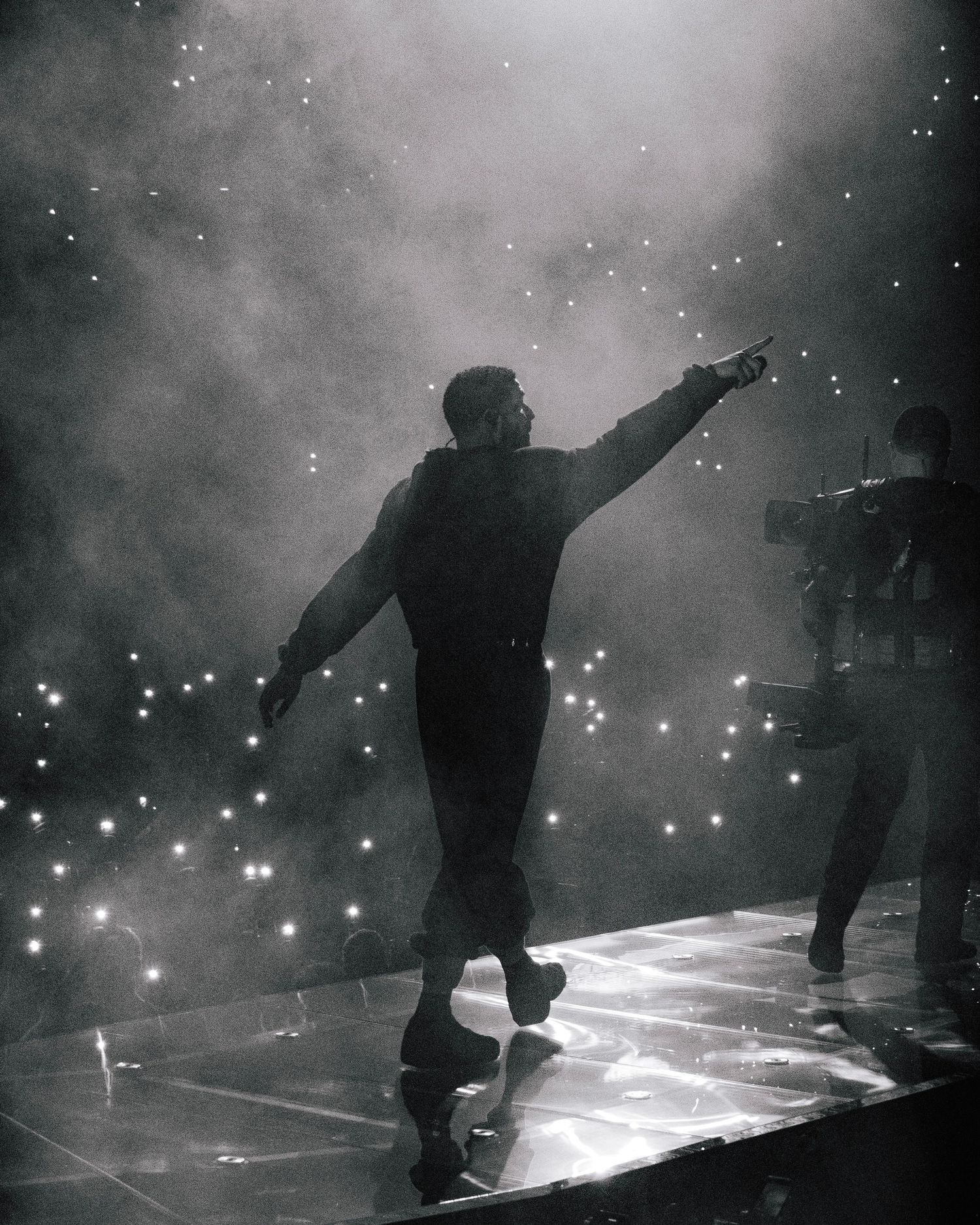 Silhouette of a performer on stage reaching upward amid fog and glittering lights in a dramatic concert moment.