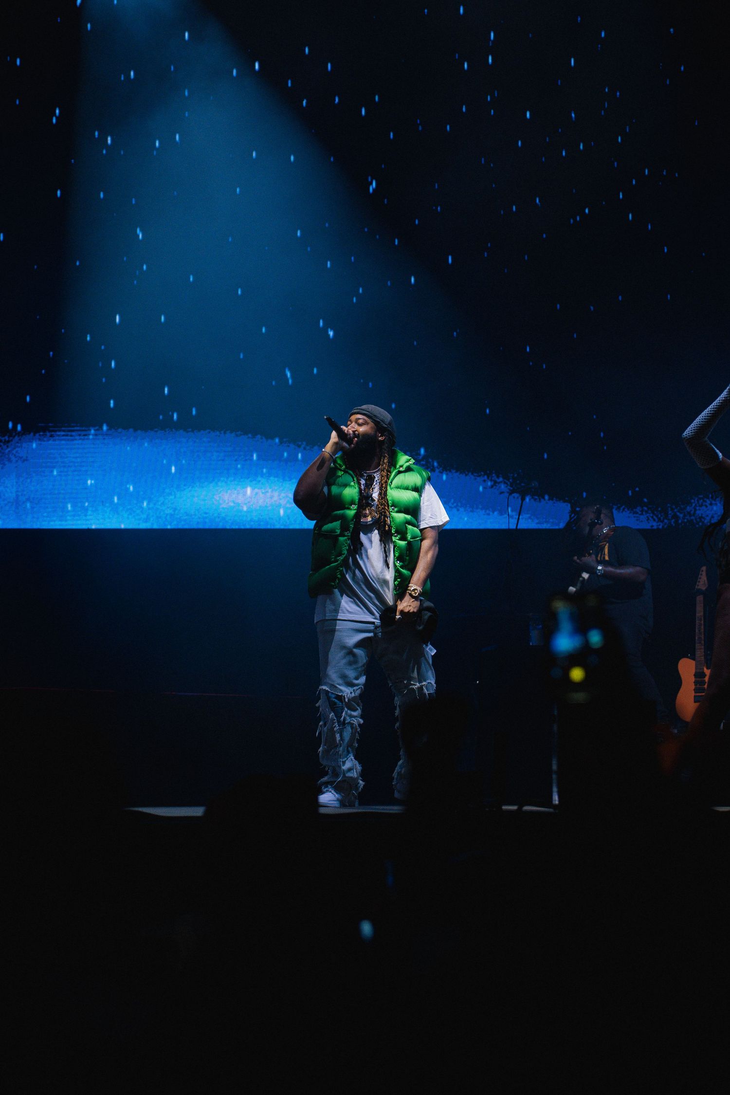 A performer in a neon green vest stands on stage under dramatic blue lighting and starry effects during a concert.