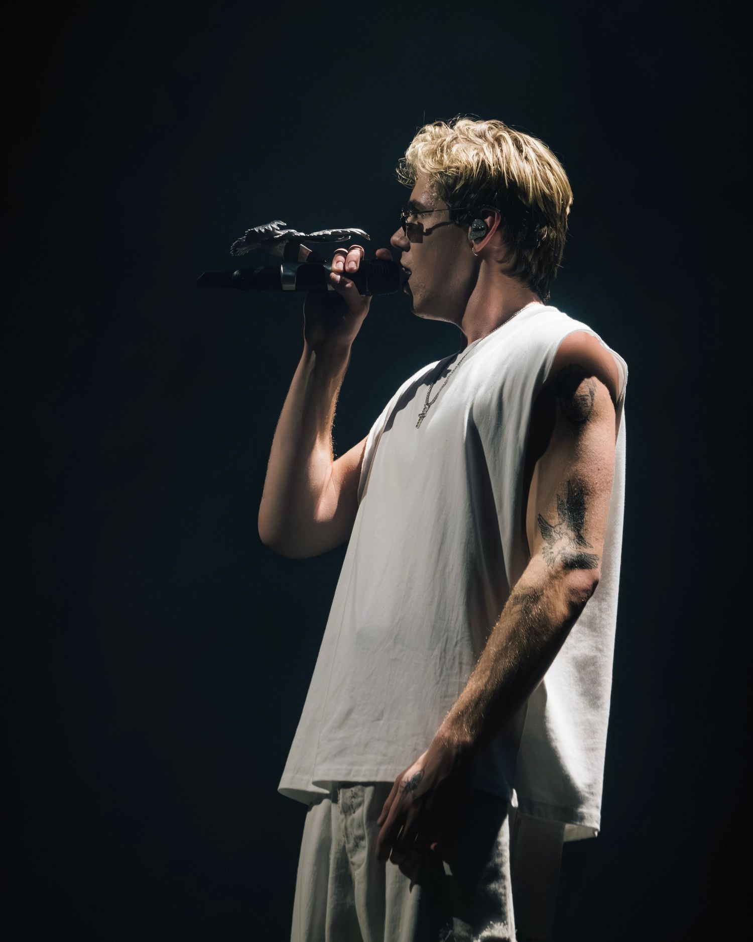 A performer in a white sleeveless shirt sings into a microphone on a darkened stage.