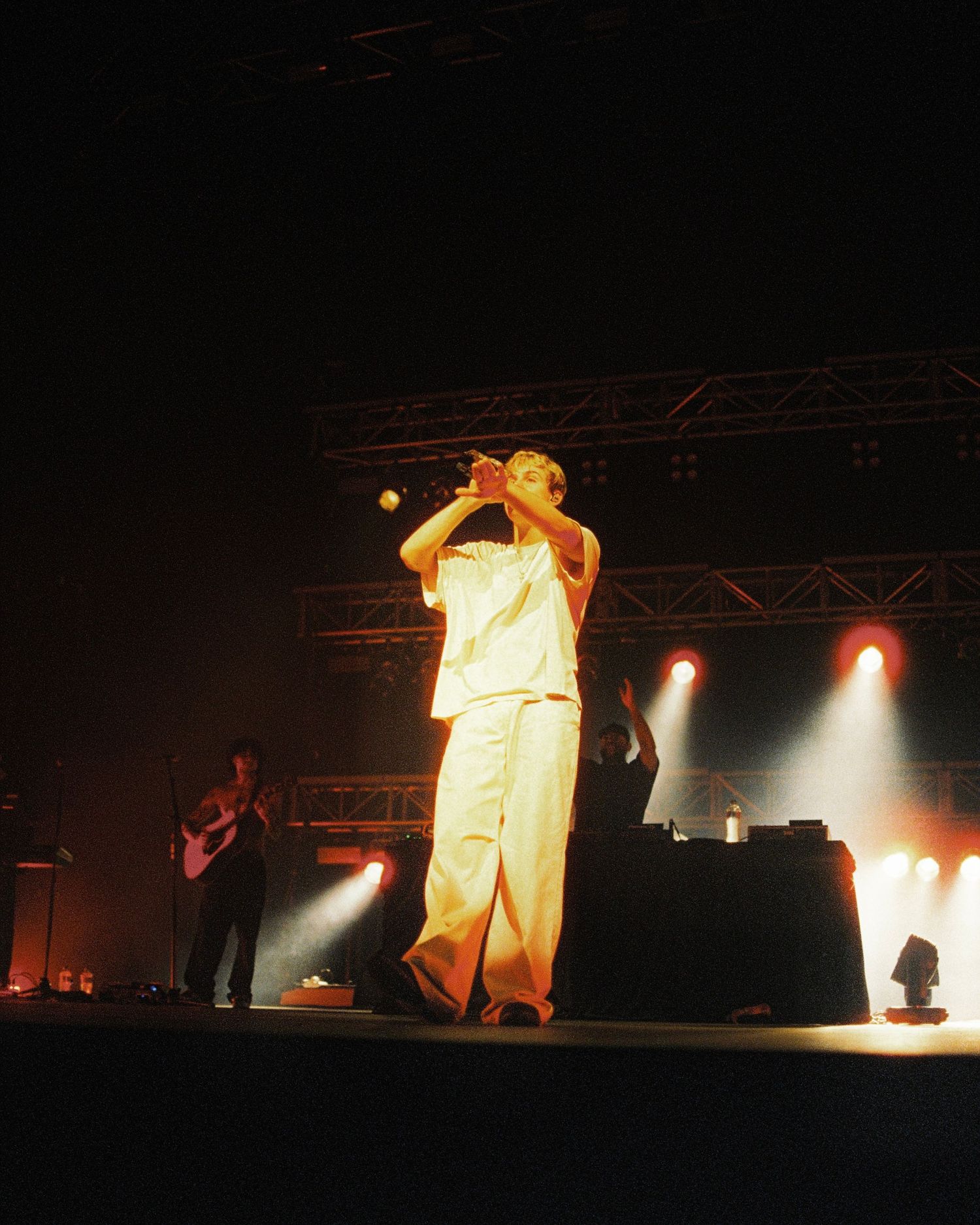 A performer in white clothing stands on stage with dramatic lighting and stage equipment visible in the background.