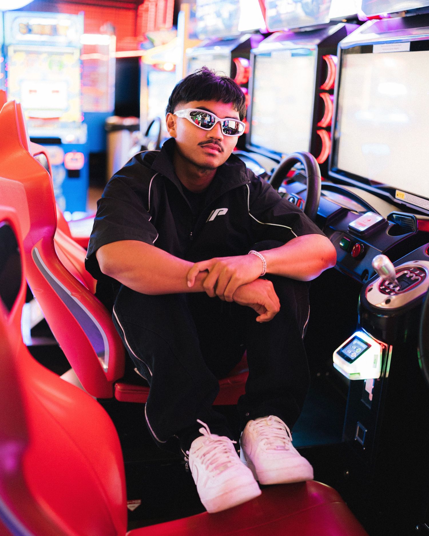 Someone sits in a red arcade seat wearing sunglasses and dark clothing against a vibrant neon-lit gaming background.