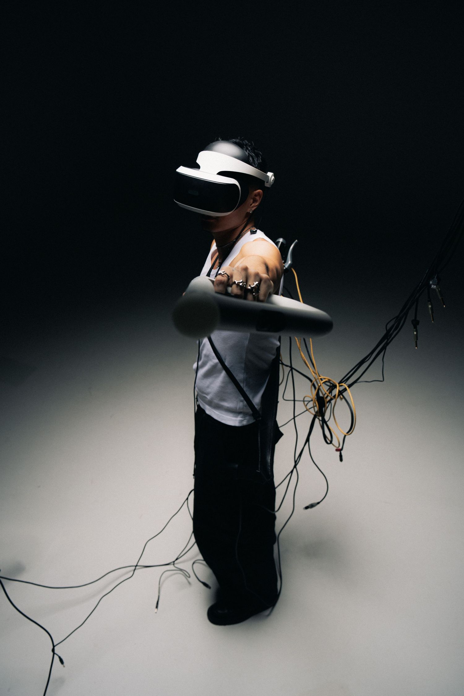 Person wearing VR headset and gray shirt stands with hanging cables against dark background.