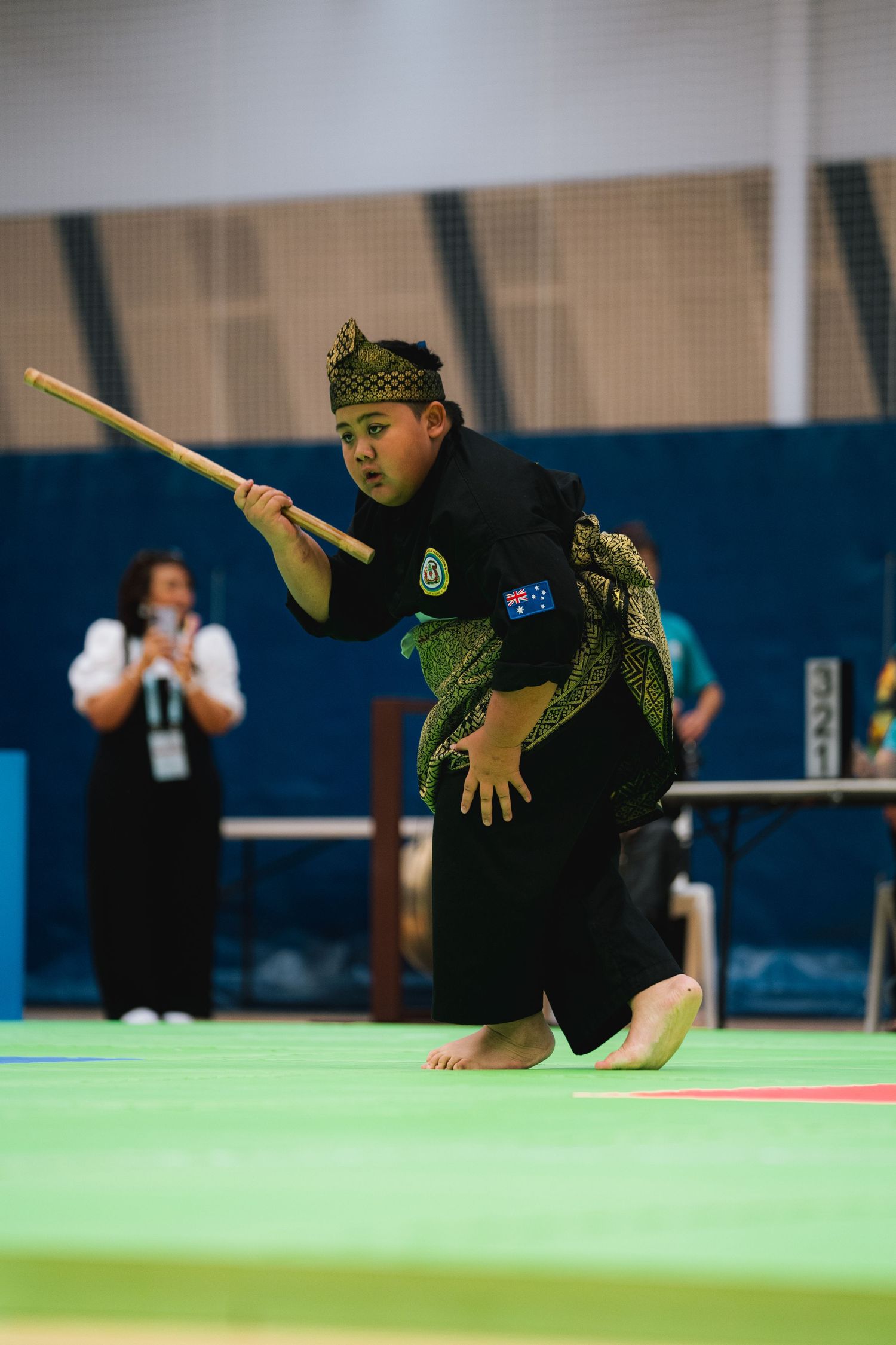 Performer in traditional martial arts costume demonstrates weapon form with a staff on green mat.
