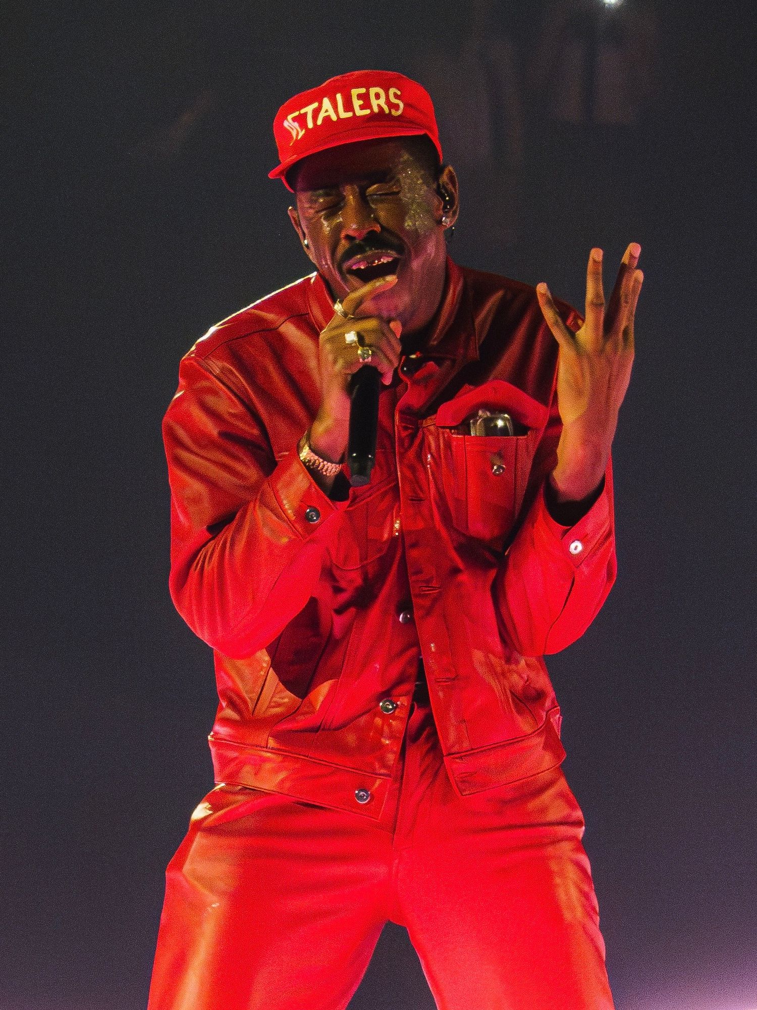 A performer in a bright red outfit strikes animated poses during what appears to be a stage performance in low lighting.