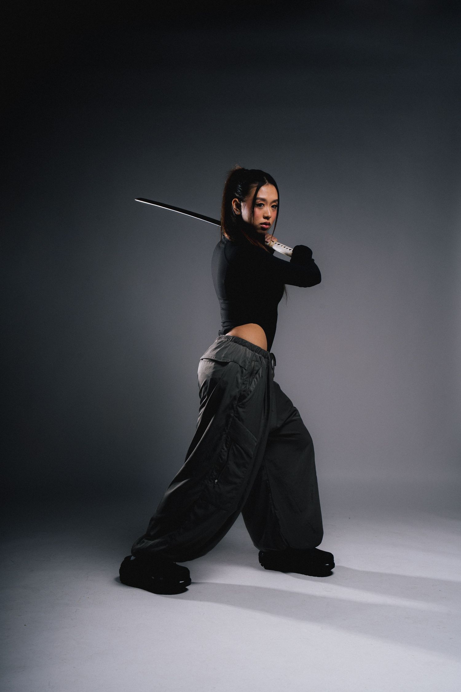 A dancer in a black crop top and baggy pants strikes a dynamic pose with a samurai sword against a dark studio backdrop.