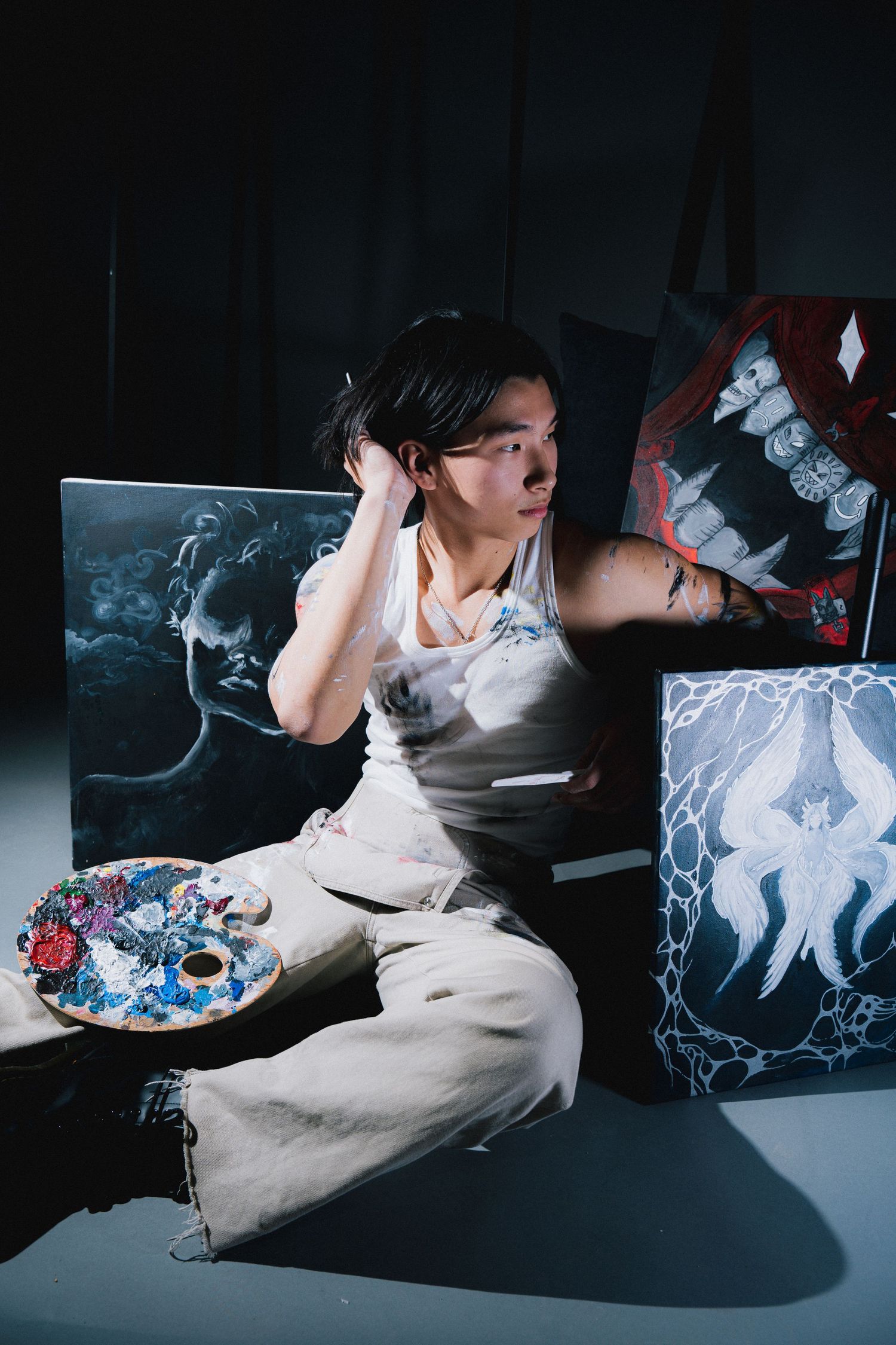 Artist in white outfit crouches with paint palette among dark moody artwork featuring ethereal smoke imagery.