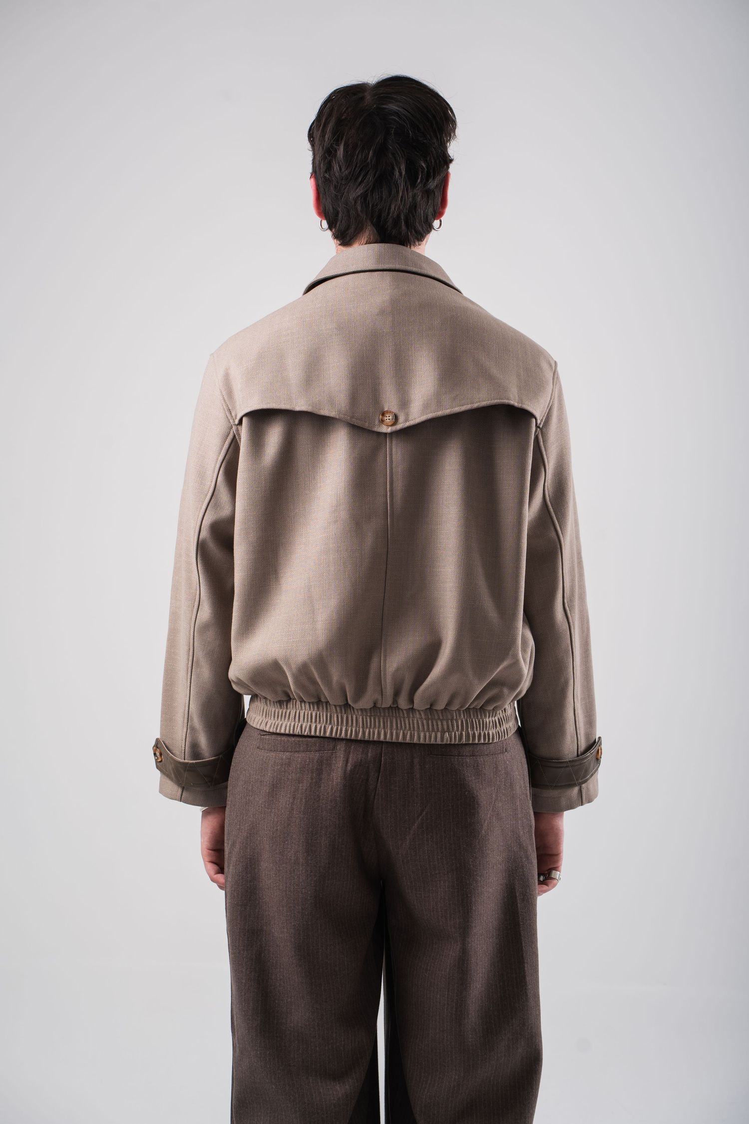 Back view of person wearing beige jacket with large collar and brown trousers against white background.