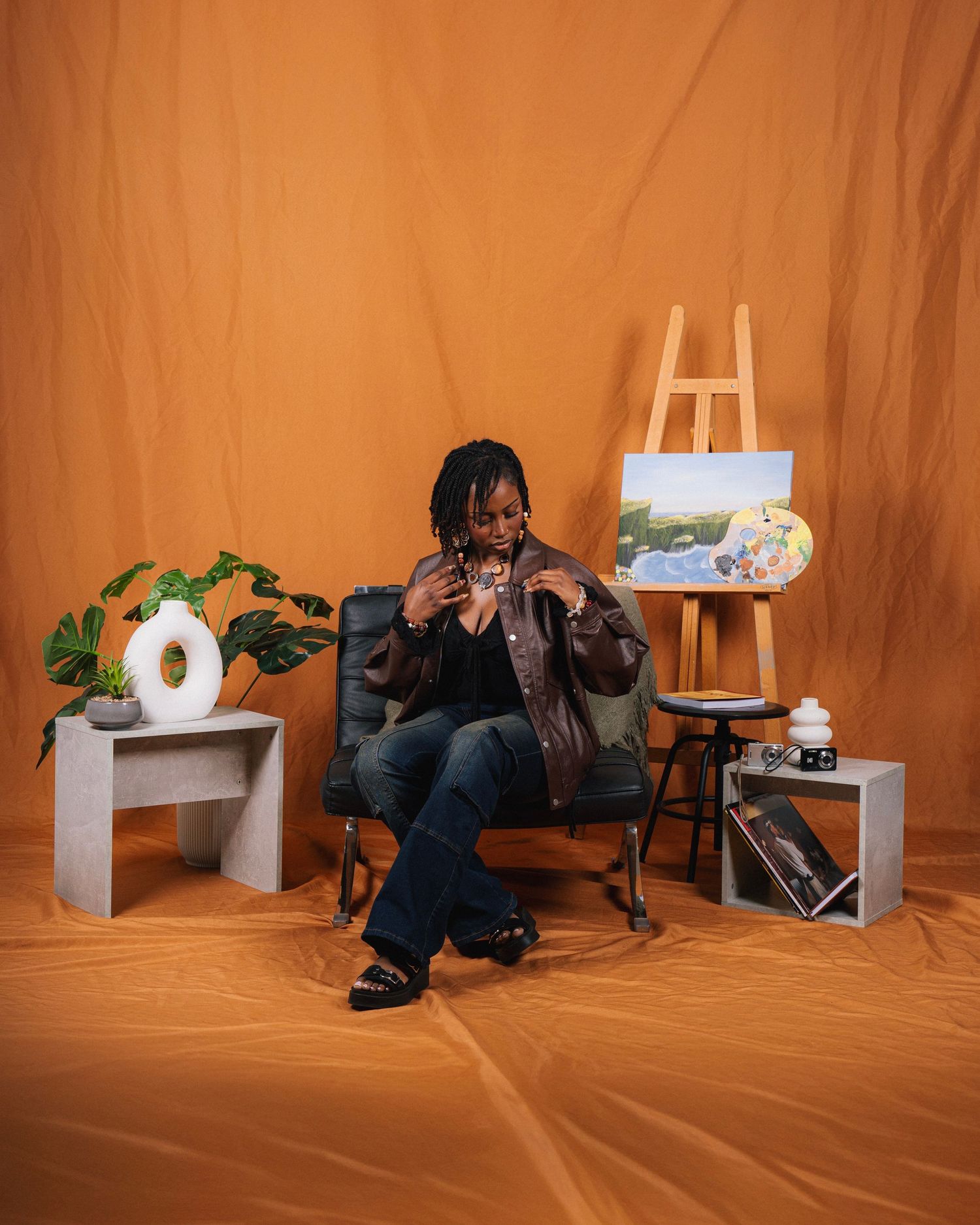 A person in a studio setting crouches near a brown backdrop with various lighting equipment and furniture arranged around them.