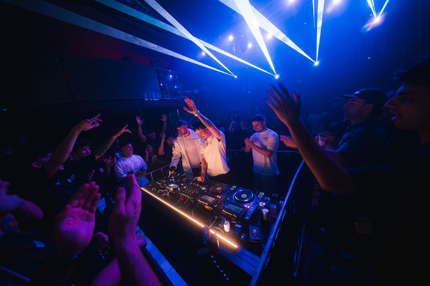 Concert crowd dancing in dark venue with blue lighting and raised hands during energetic performance.